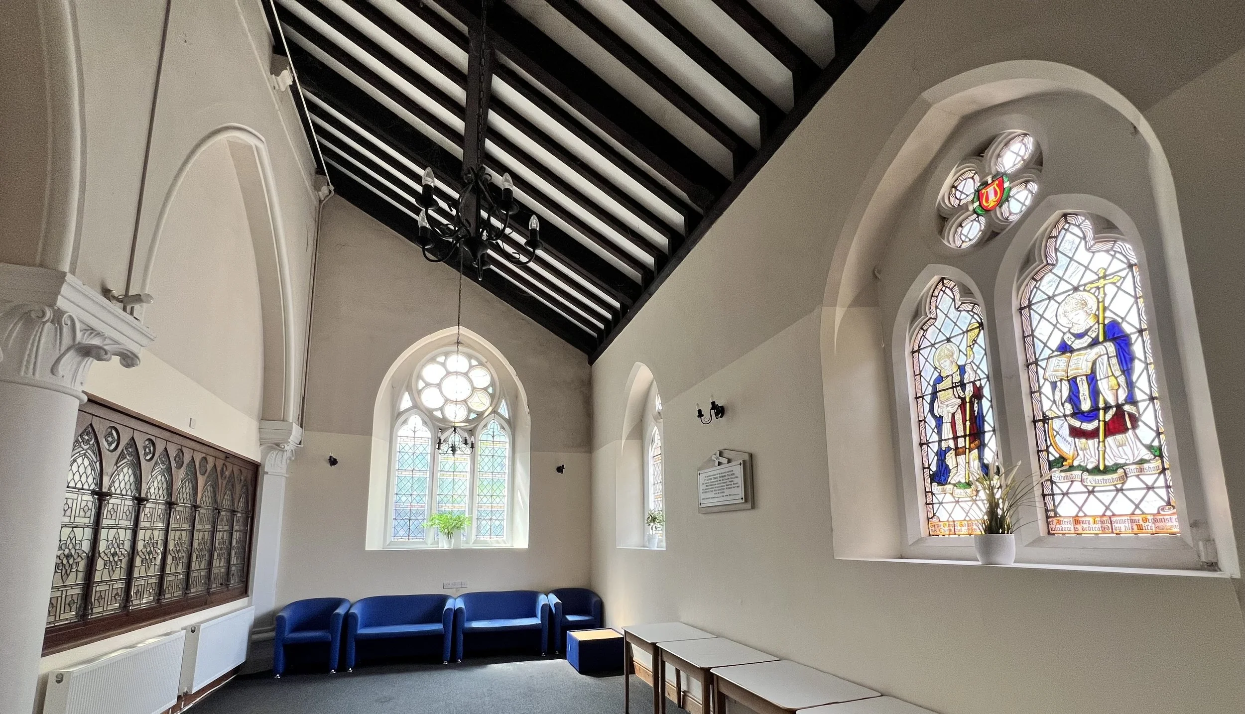 Interior of a church with stained glass windows, a black chandelier, blue seating, and a white wall with decorative columns.