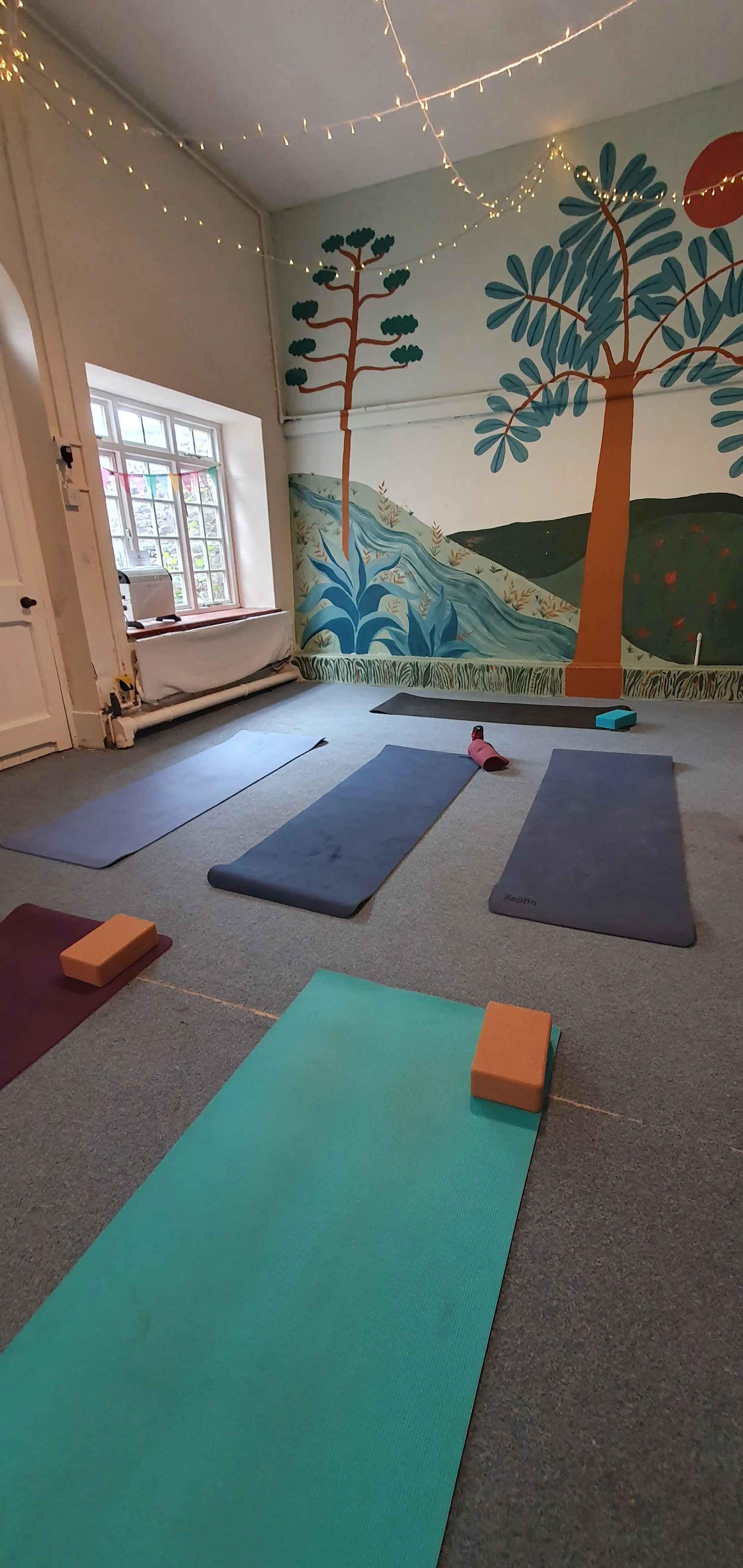 Interior of a yoga studio with yoga mats, blocks, a blue foam roller, and a mural of trees and a river on the wall, illuminated by string lights.