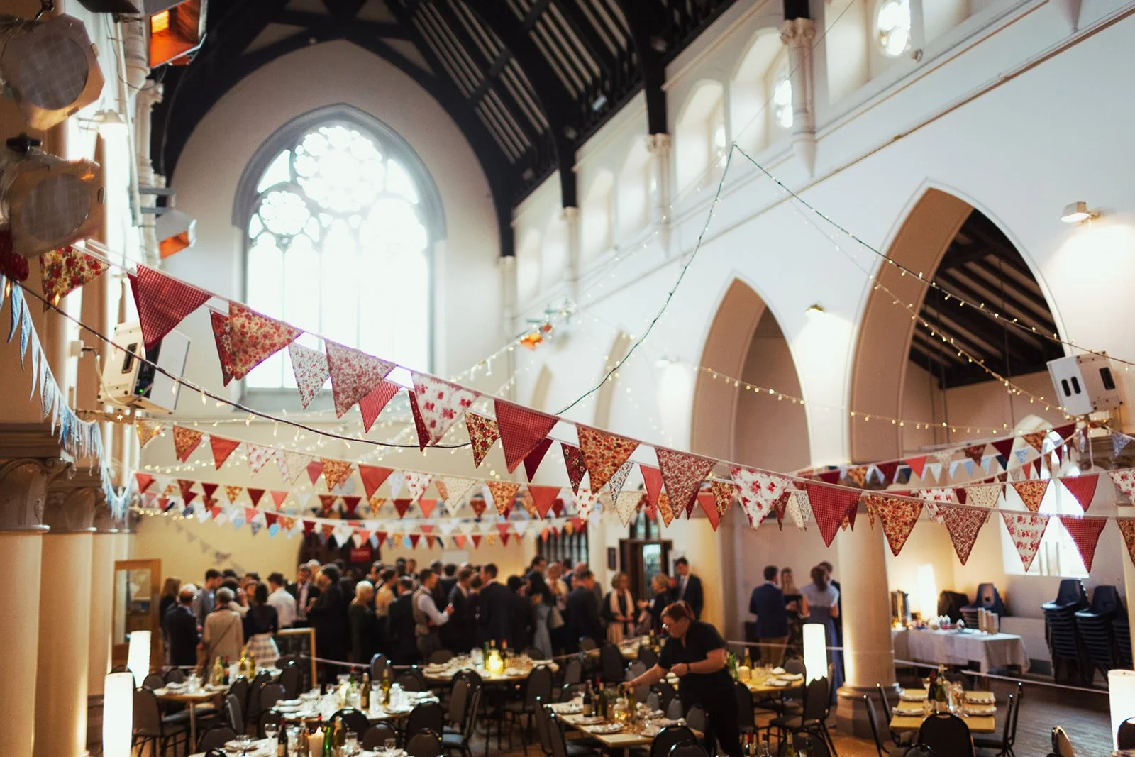 Indoor event space decorated with red and white bunting and string lights, with tables set for a celebration and people mingling.