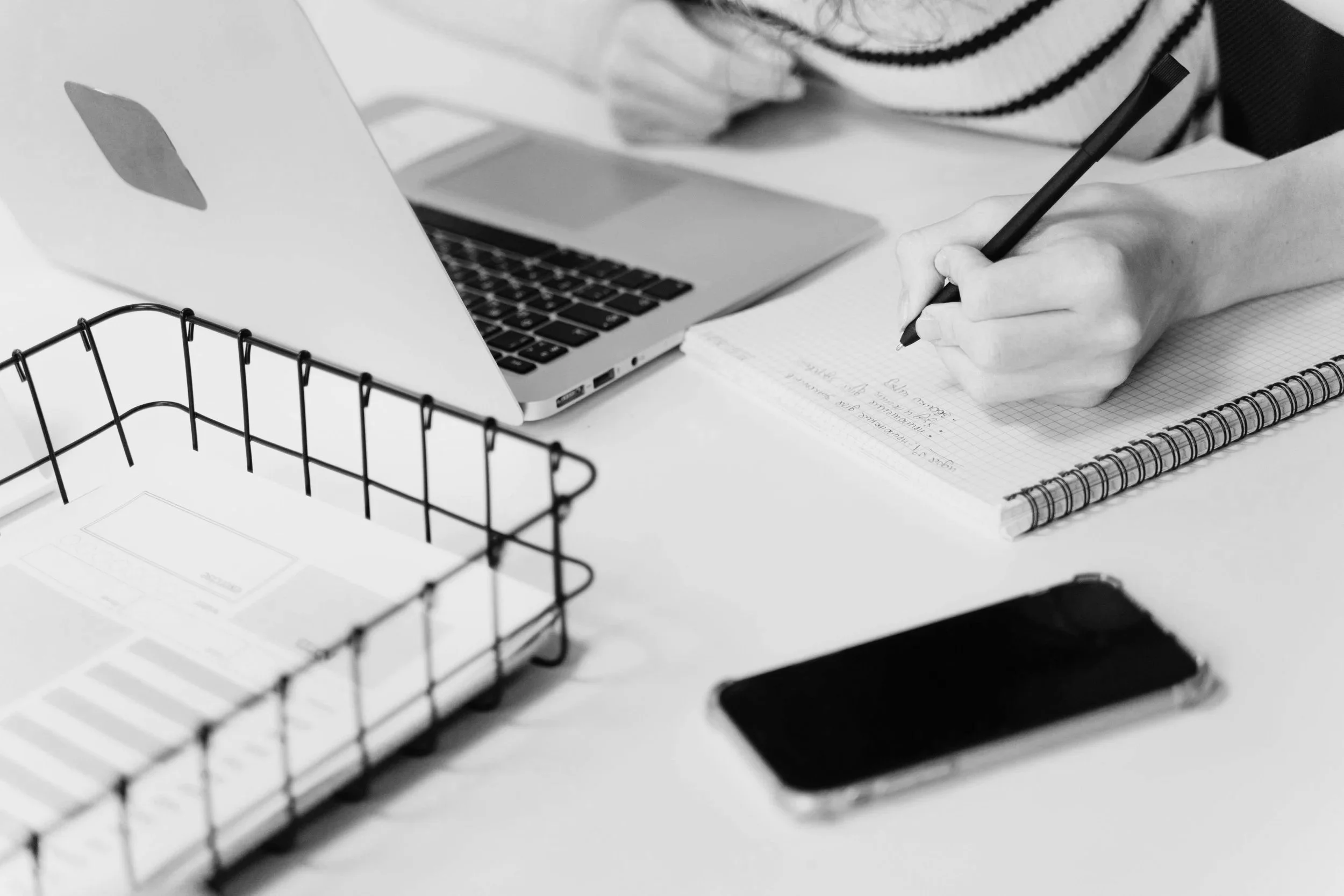 Eine Person schreibt in ein Notizbuch, neben einem Laptop, einem Smartphone und einer Aktenablage auf einem weißen Tisch.