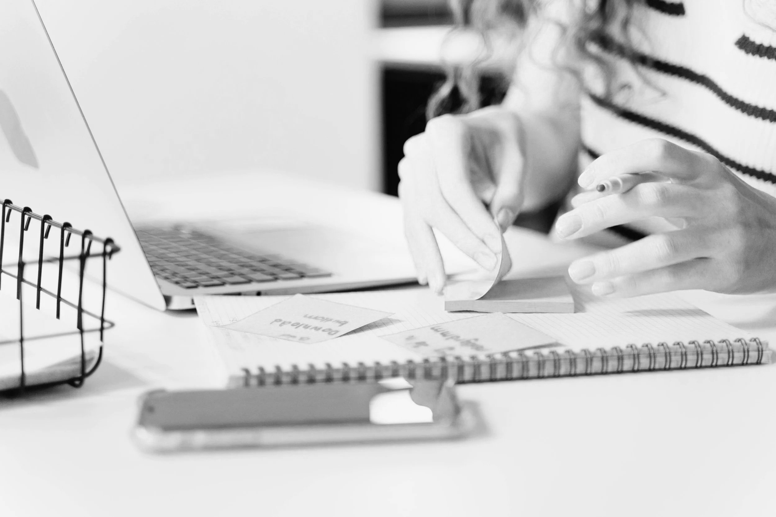 Schreibtisch mit Laptop, Notizbüchern, Notizzetteln und einem Handy, Person schreibt Notizen in der Hand, Schwarz-Weiß-Foto.