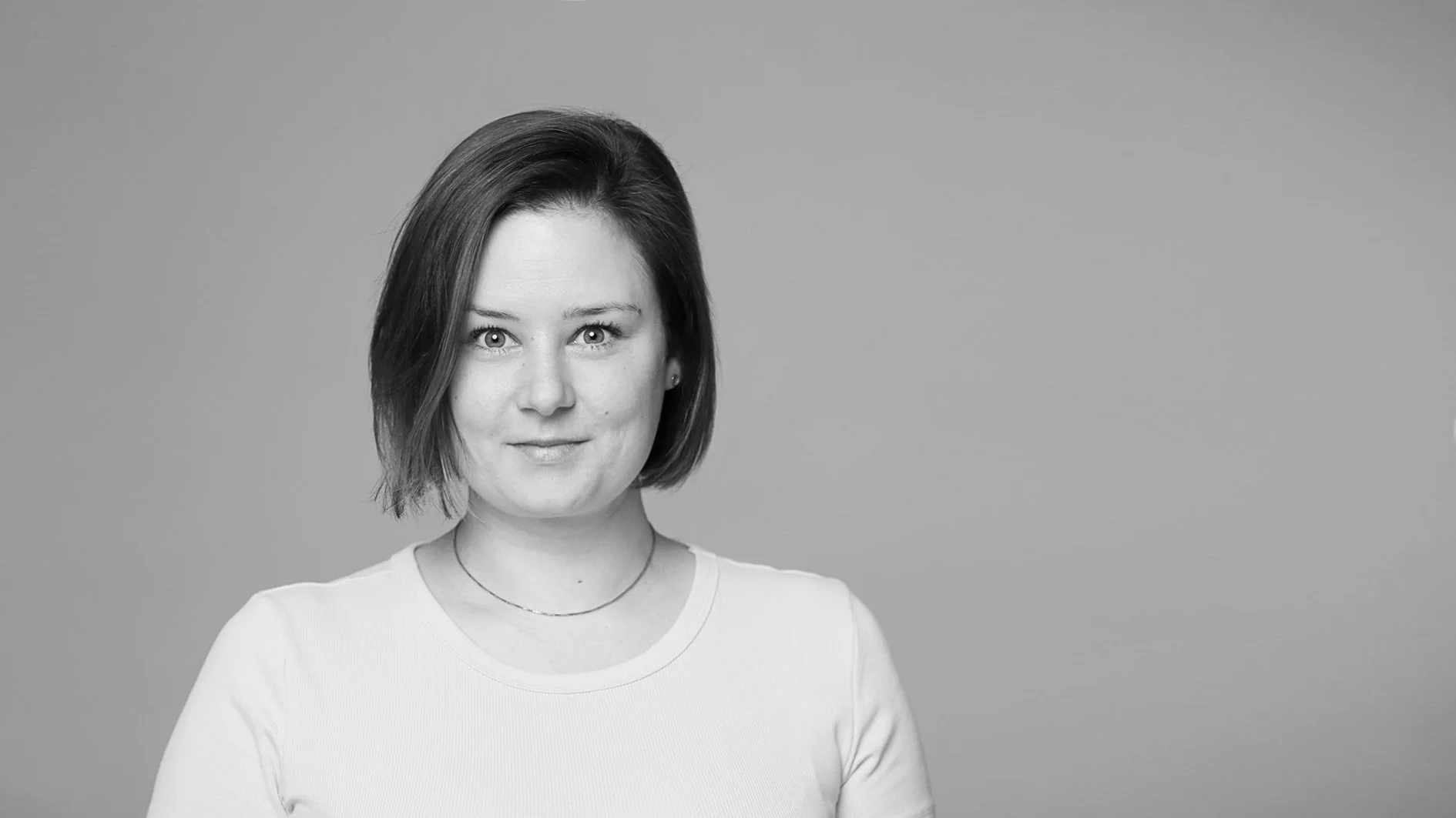 Schwarz-weiß Porträt einer Frau mit kurzen braunen Haaren, trug ein weißes T-Shirt, blickt direkt in die Kamera, neutraler Hintergrund.
