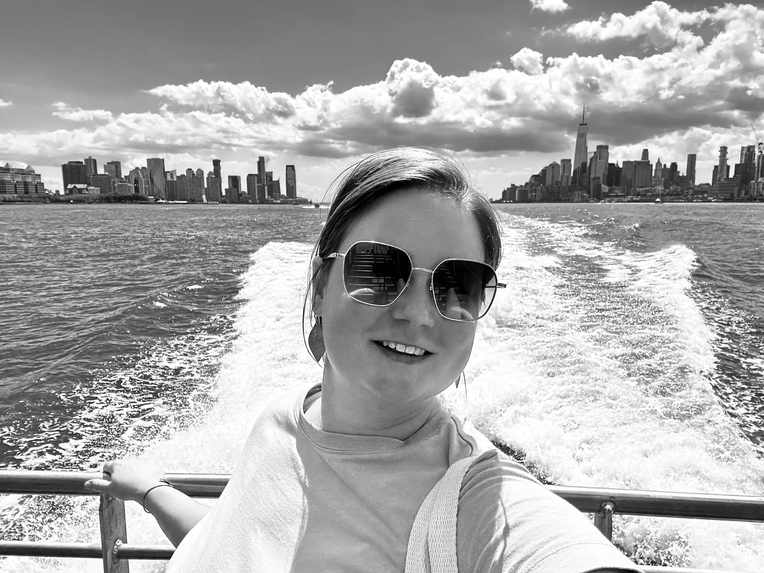 Lächelnde Frau mit Sonnenbrille macht Selfie auf Boot mit Stadt skyline im Hintergrund, New York City, Himmel mit Wolken, Wasser mit Wasserlinie im Wasser