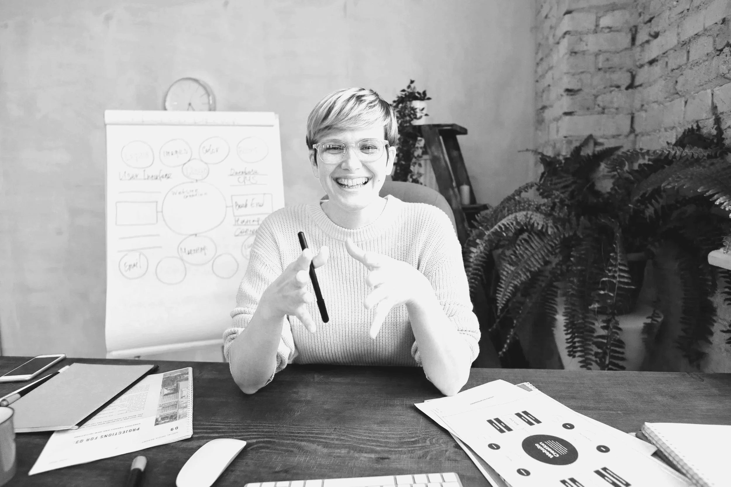 Eine lachende Frau sitzt an einem Schreibtisch mit Papieren, einem Tablet, einem Smartphone und einem Marker, im Hintergrund eine Tafel mit Diagrammen, Pflanzen und Bücher.