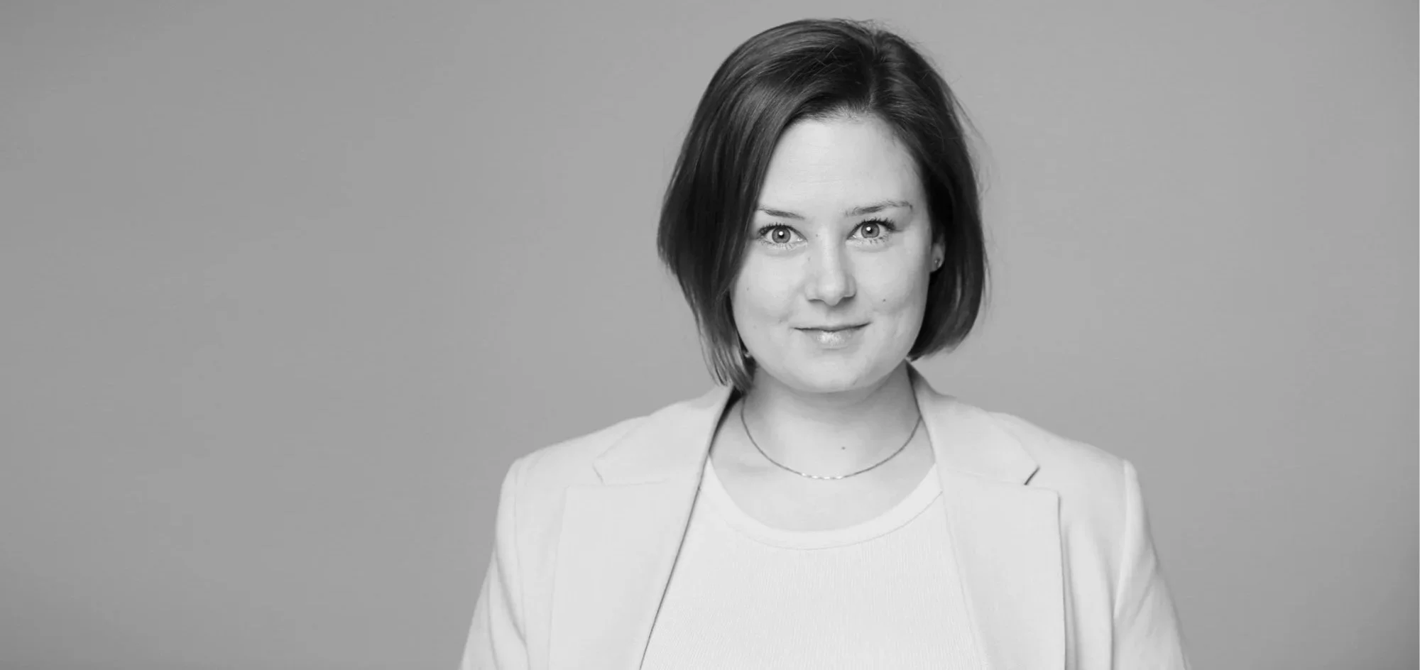 Schwarz-weiß-Fotografie einer jungen Frau mit kurzen Haaren, die einen weißen Blazer trägt, vor grauem Hintergrund.