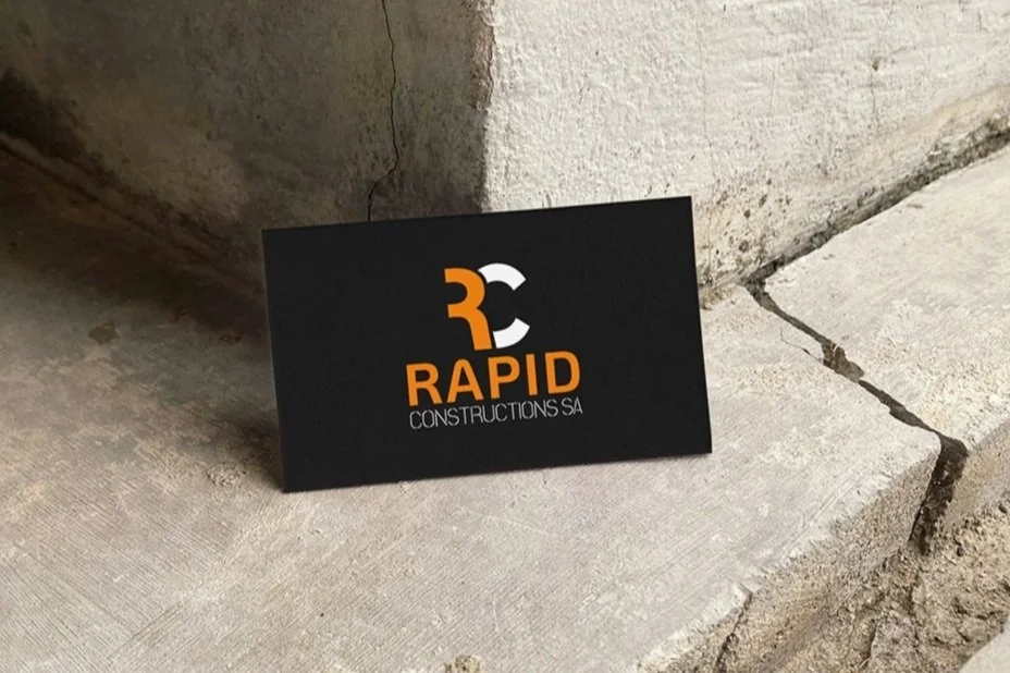 Business card for Rapid Constructions SA, black background with orange and white logo and text, placed on concrete surface near a wall.