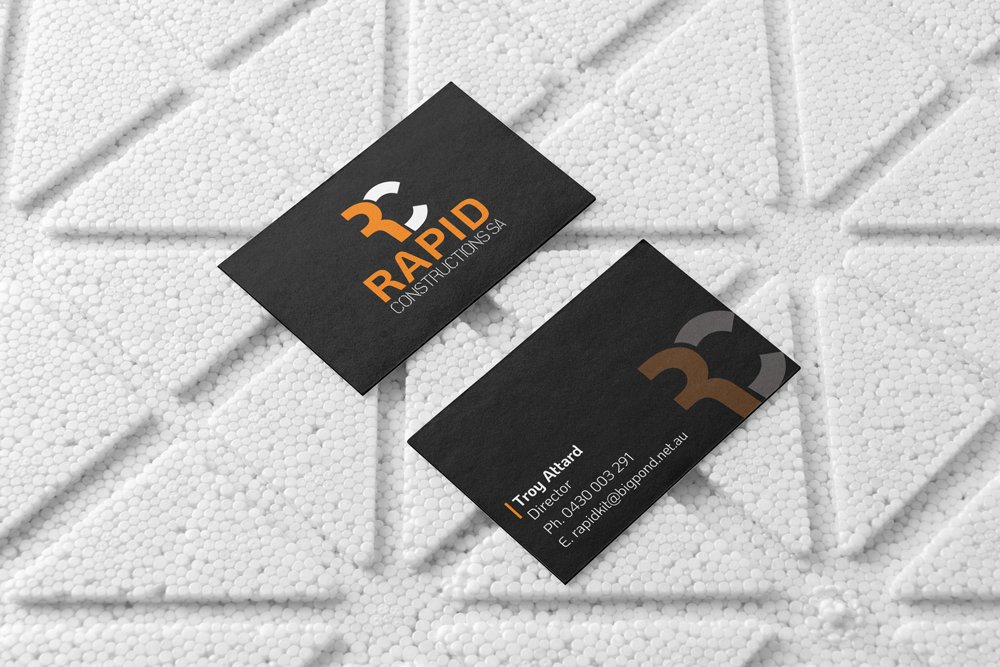 Black business cards with the logo and contact information of Rapid Construction SA placed on a white textured surface.