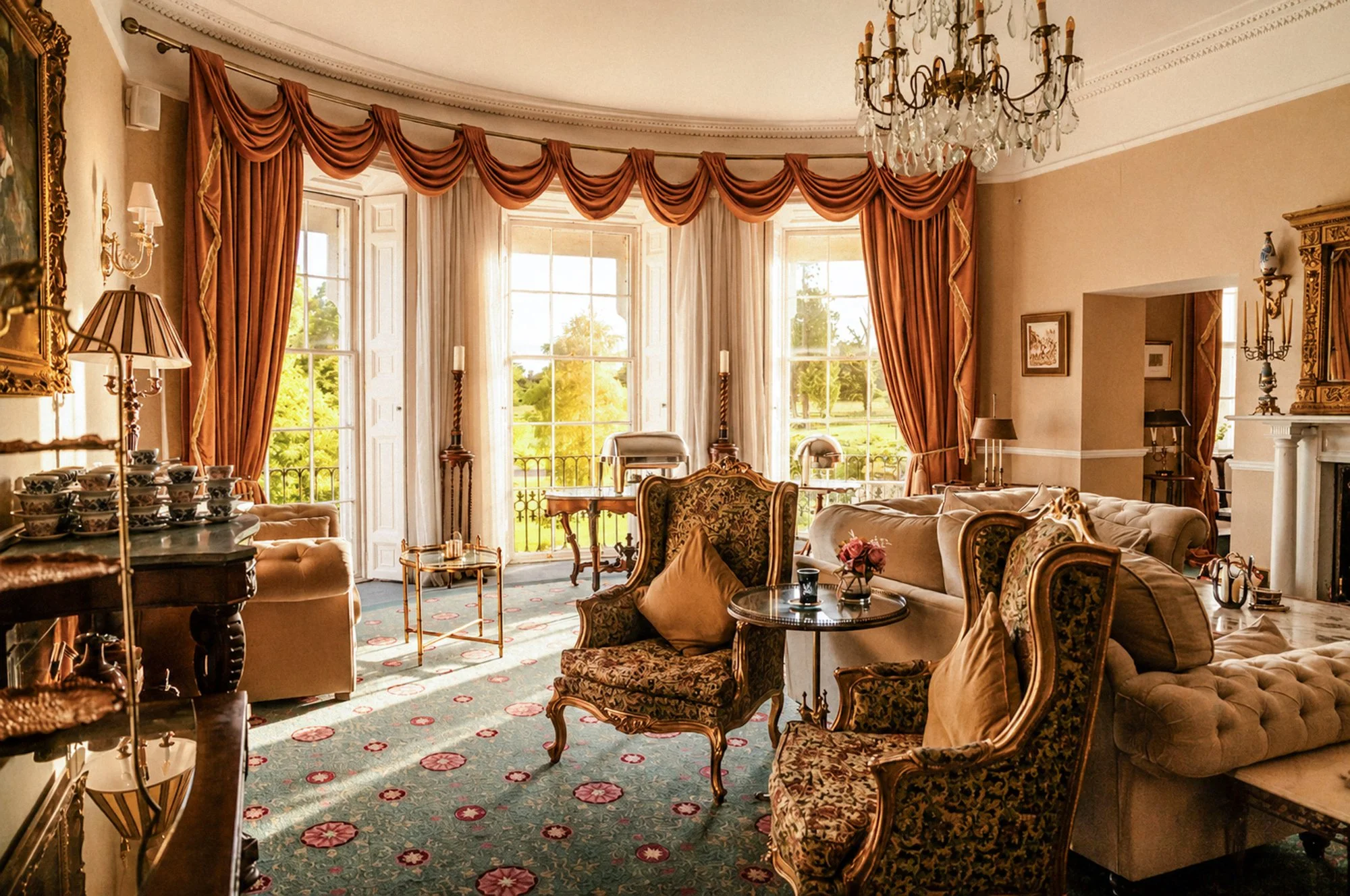 Elegant living room with large bay windows, ornate drapes, antique furniture, a chandelier, and decorative items.