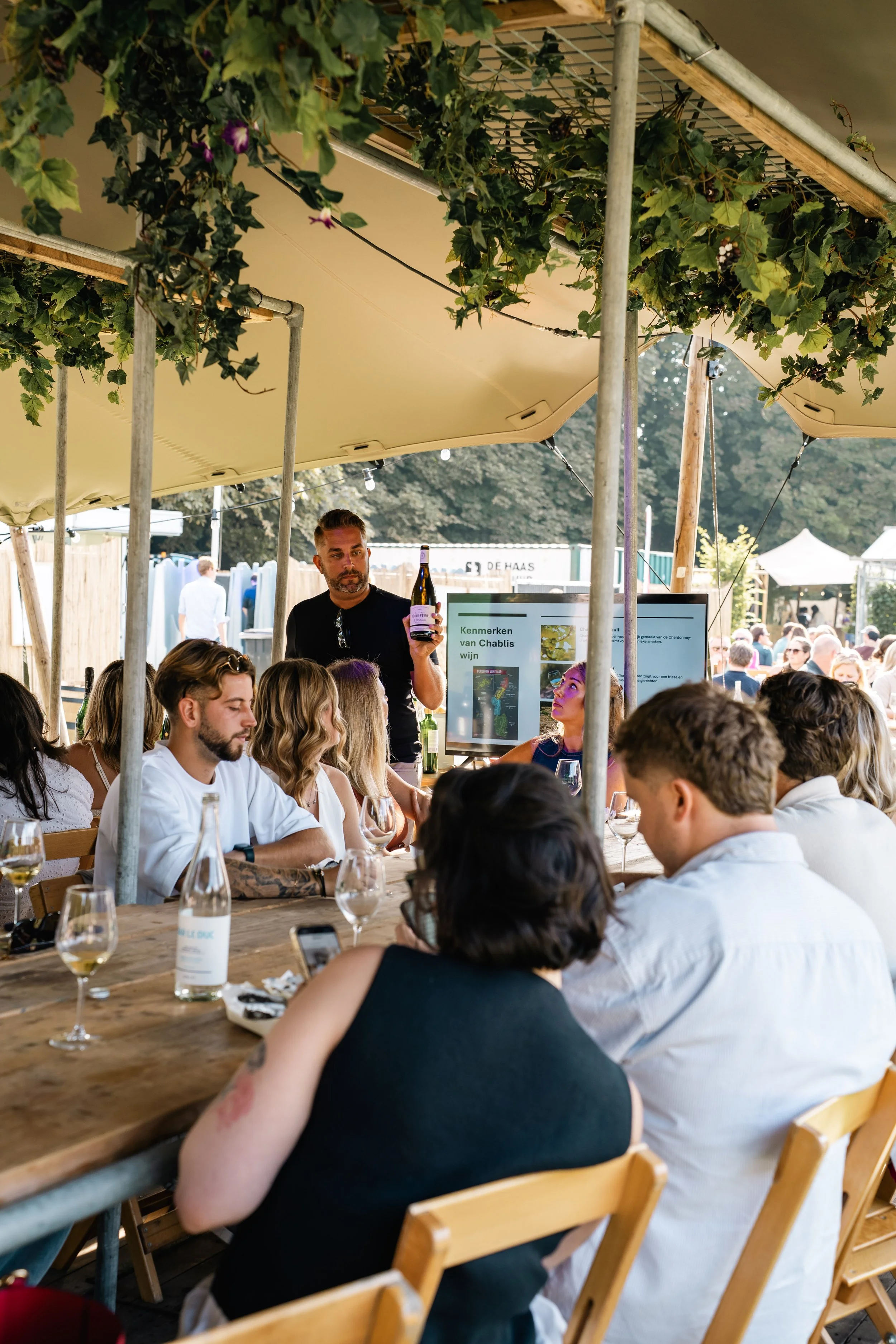 Mensen zitten aan een lange houten tafel onder een tent met groene bladeren en verlichting, met een man die wijn vasthoudt.