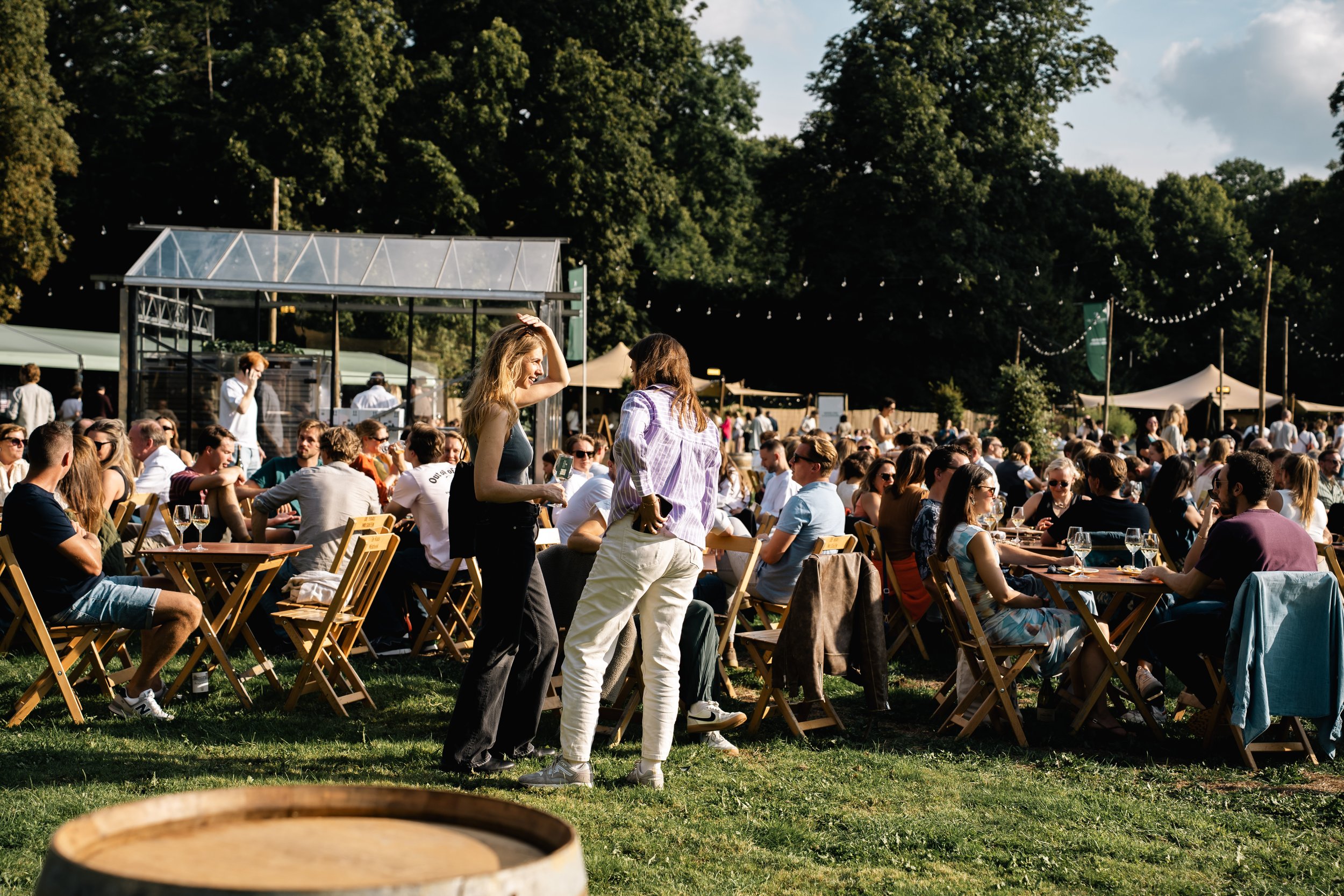 Een groot buitenevenement met veel mensen die aan houten tafels zitten en genieten in de zon. In de achtergrond zijn tenten, een foodtruck en stevige bomen zichtbaar, met blauwe lucht en enkele wolken.