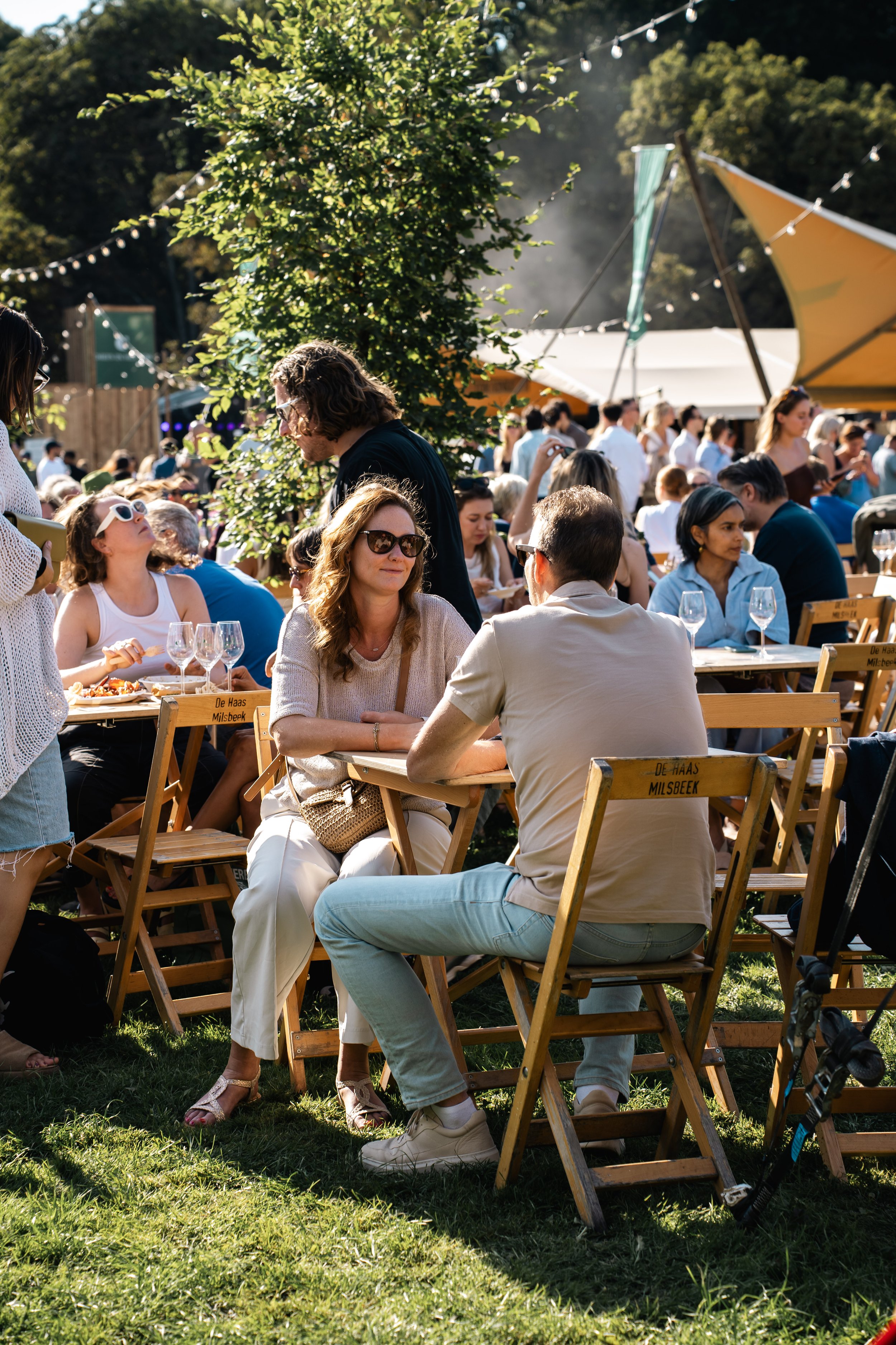 Mensen zitten aan houten tafels onder een grote tent op een zomers evenement, groene bomen en kerstverlichting in de achtergrond.