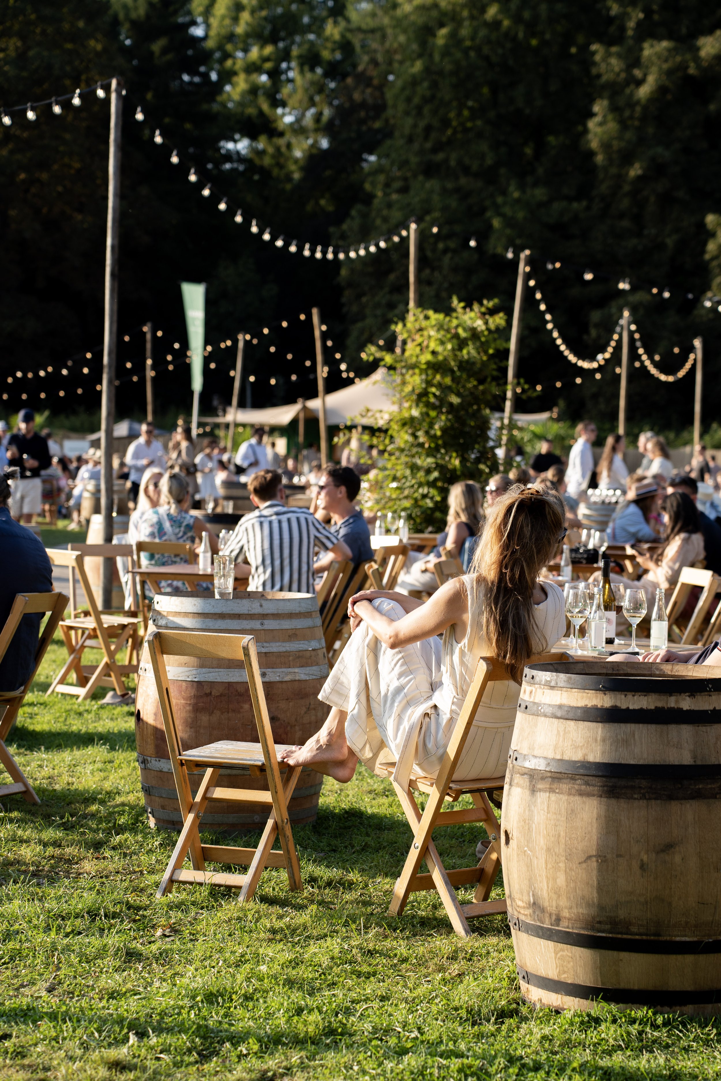 Mensen zitten buiten op houten tafels en stoelen, drinken wijn, onder een boom met lichtslingers, bij een evenement op een grasveld tijdens de zomeravond.