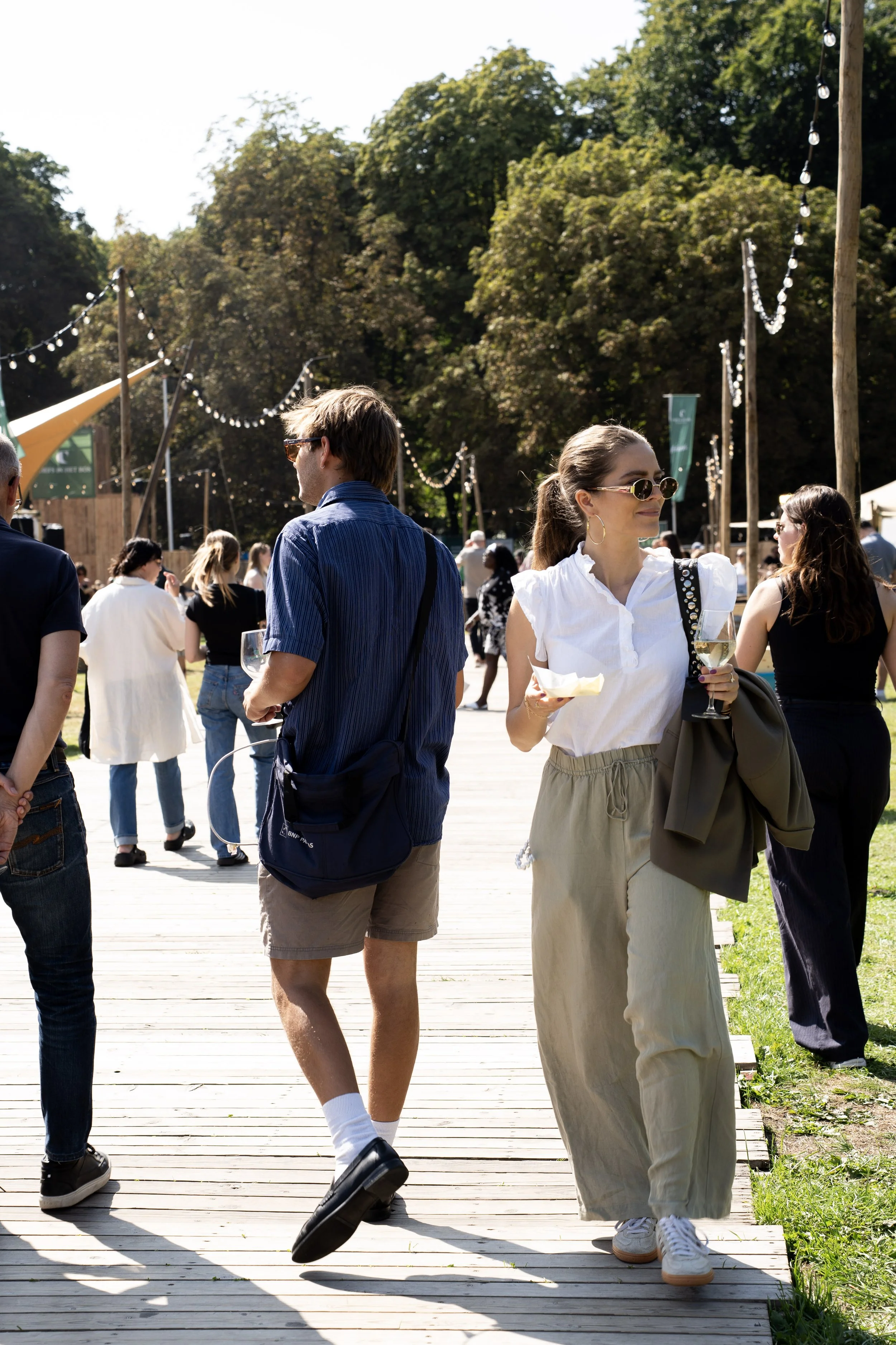 Mensen lopen op een houten wandelpad in een park tijdens een zonnige dag, sommige mensen drinken wijn, andere praten en genieten van het buiten zijn.