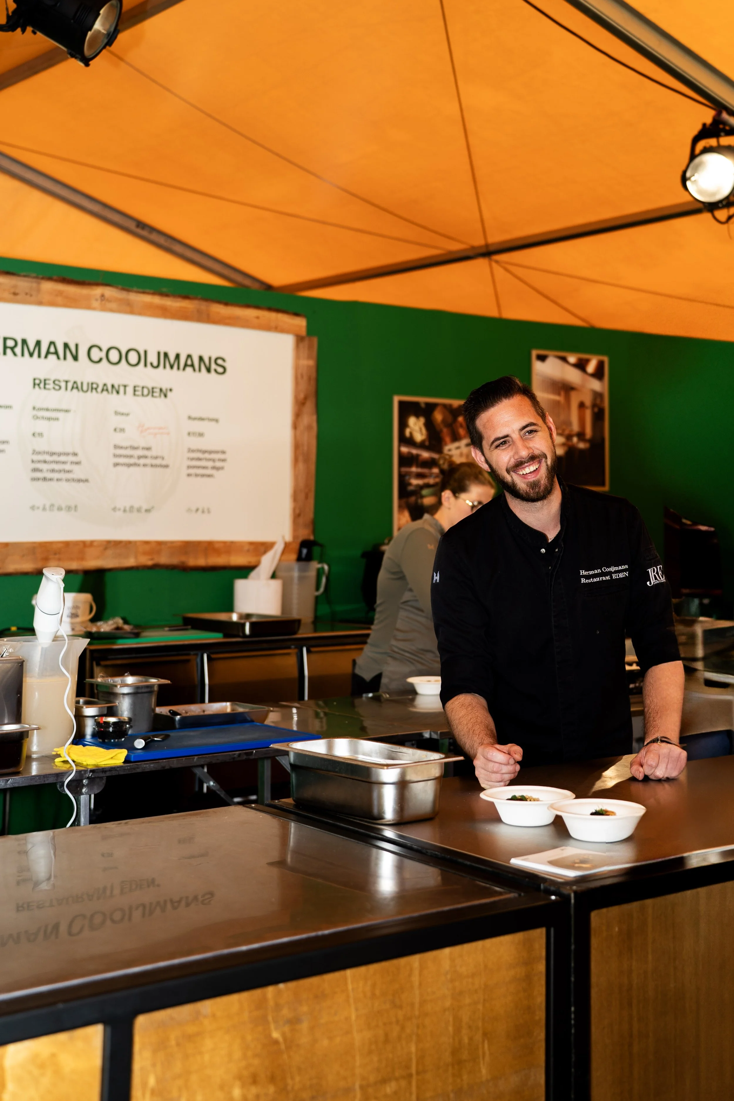 Vrolijke chef in zwart uniform achter een houten keukenblad met twee kleine gerechten, in een groene tentenhal met een geel dak.