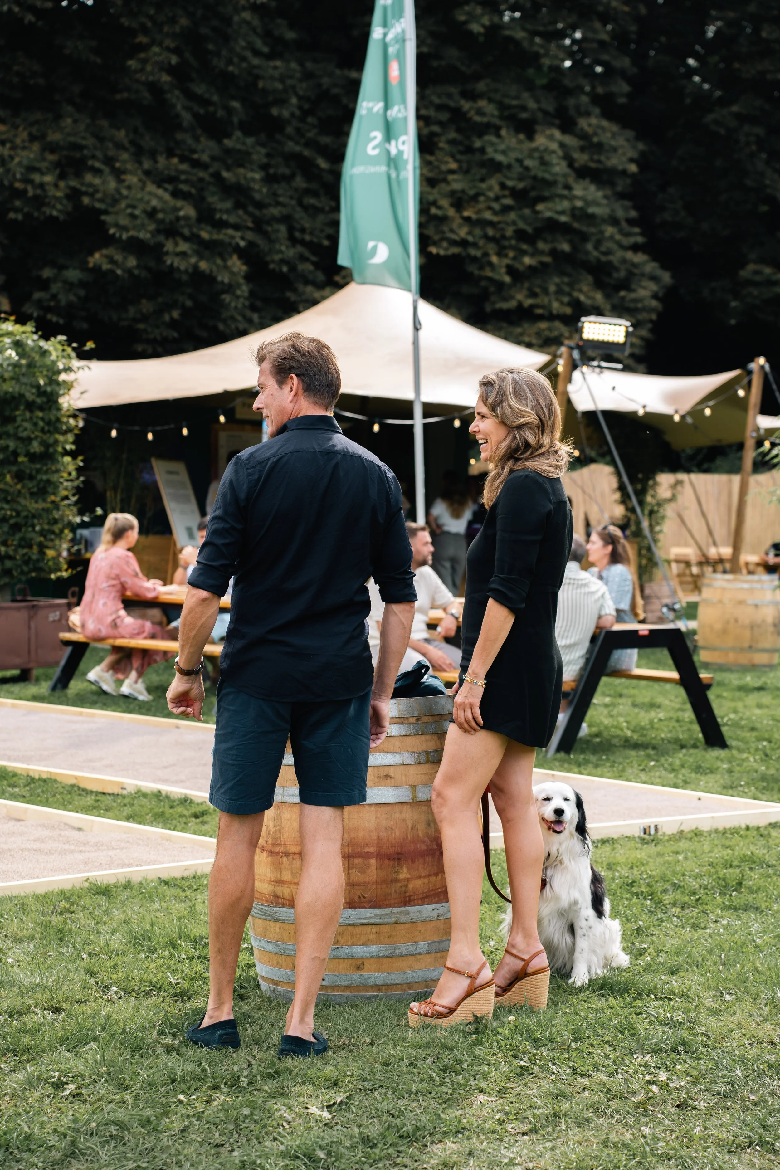 Twee mensen praten buiten in een park, een vrouw met een hond en een man, met een tent en een vlag op de achtergrond.