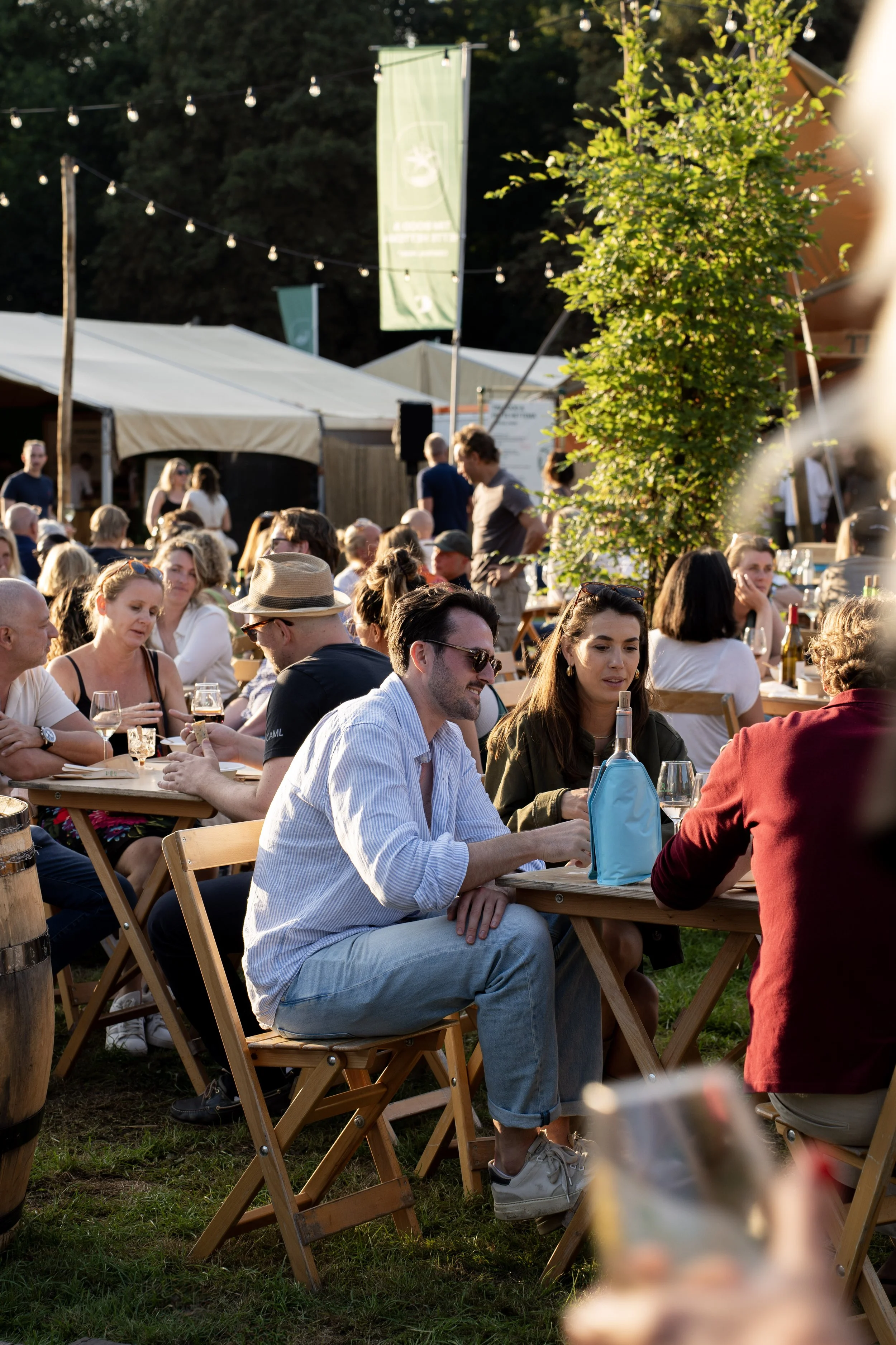 Een groep mensen zit buiten aan lange houten tafels tijdens een zomers evenement met tenten en lantaarns, genietend van eten en drinken.