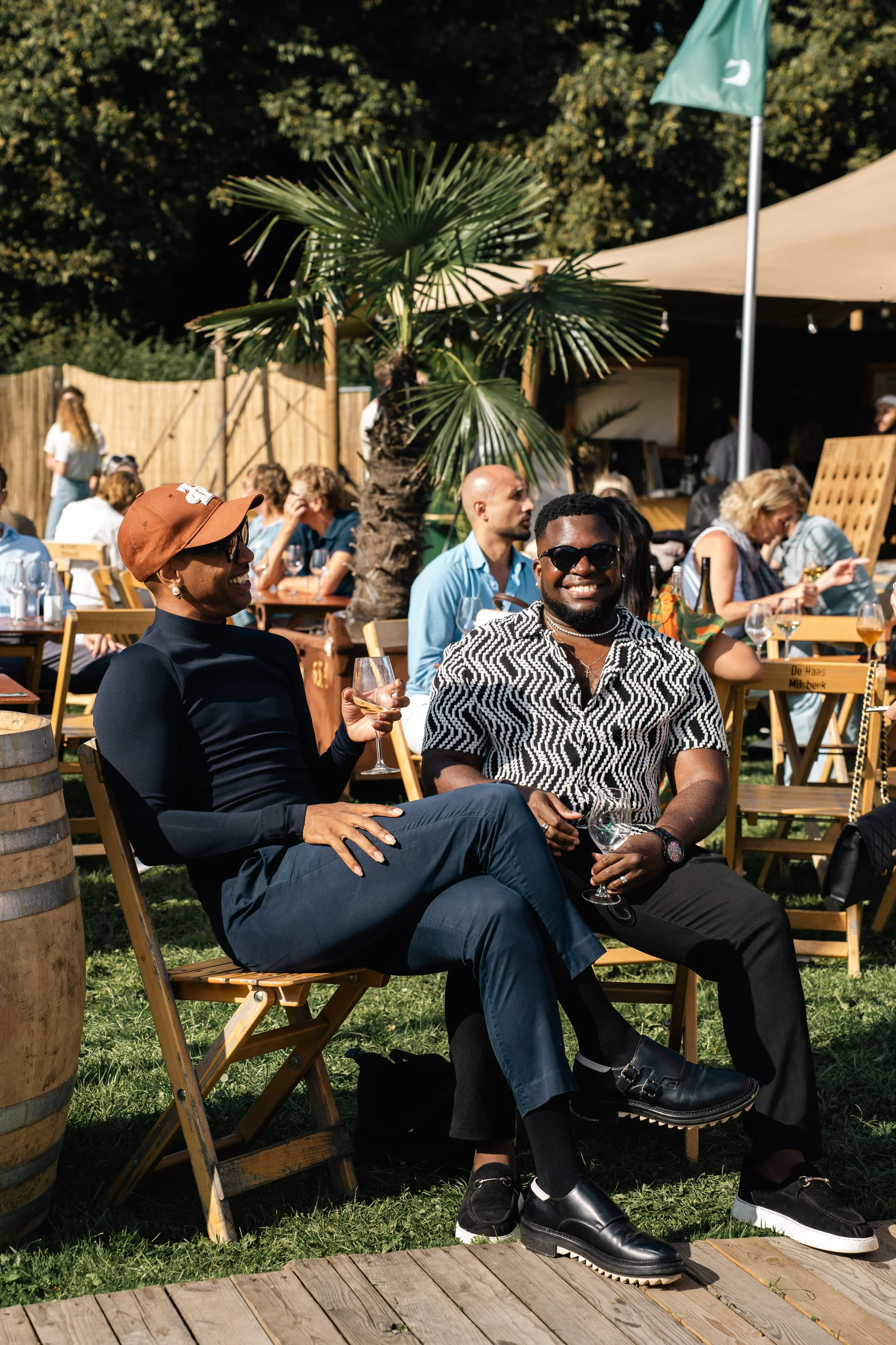 Twee mensen zitten op houten stoelen buiten, lachen en drinken wijn op een zomerdag, omgeven door andere gasten en een palmboom, met een tent en een vlag op de achtergrond.