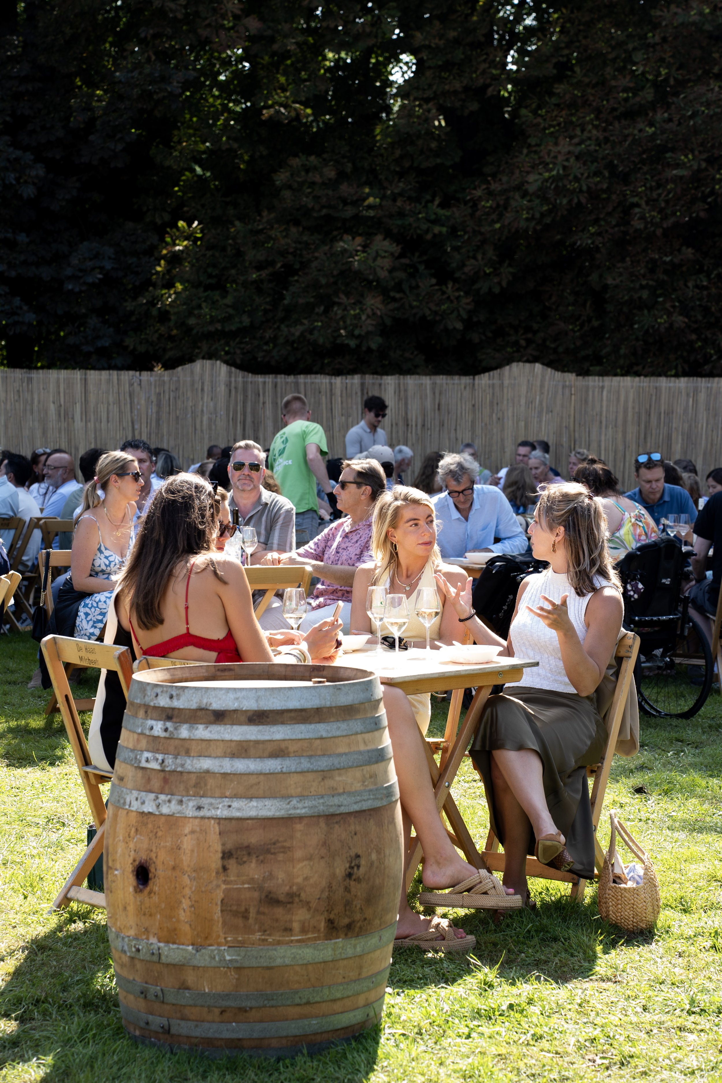 Mensen zitten aan een lange tafel in een tuin, genietend van een zomerse dag. In de voorgrond staat een grote houten vat.