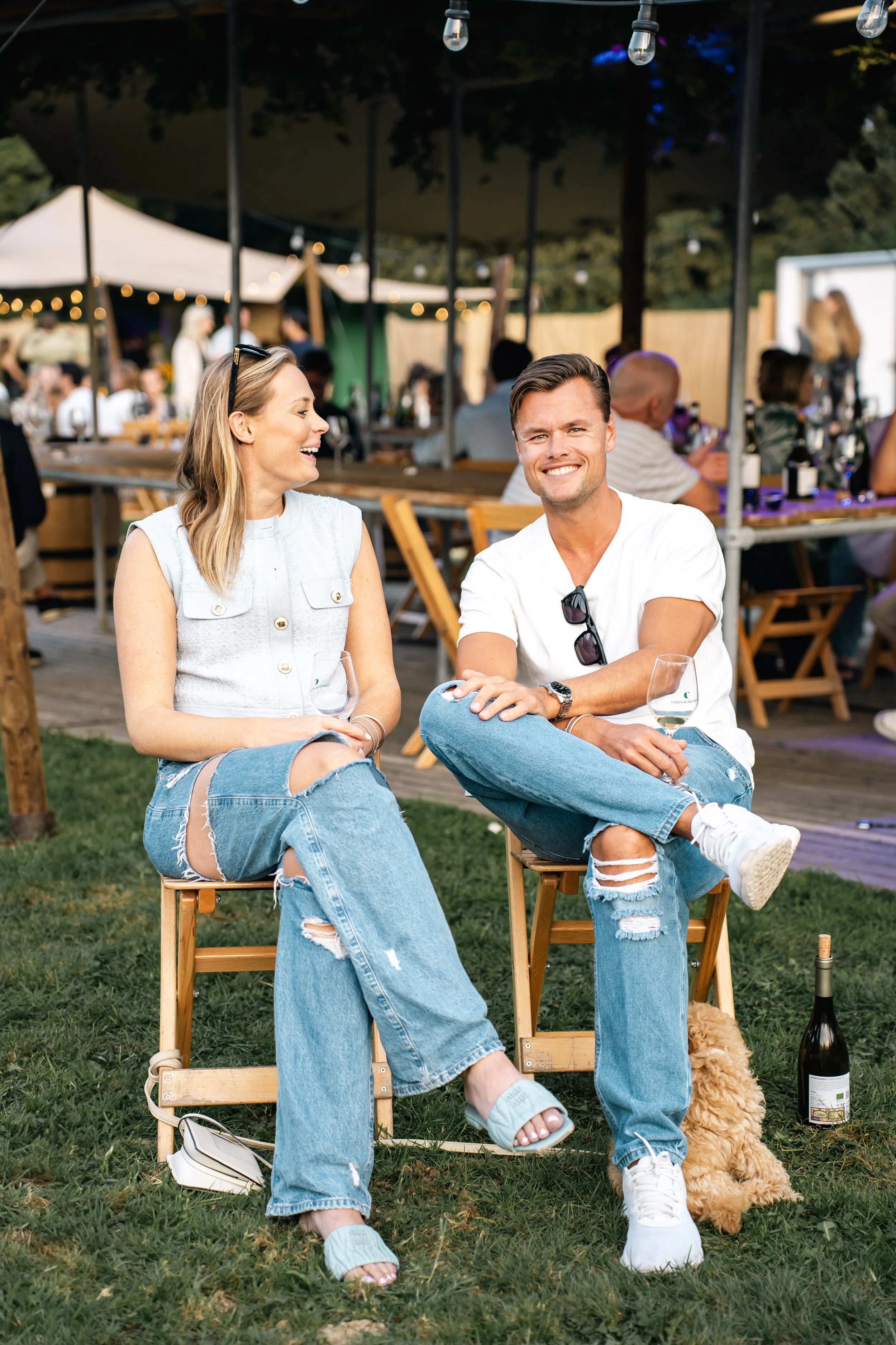 Twee lachende mensen zitten op stoelen op een grasveld tijdens een buitenfeest, met een menigte op de achtergrond onder een overkapping en feestverlichting, een wijnfles en een hond naast hen.