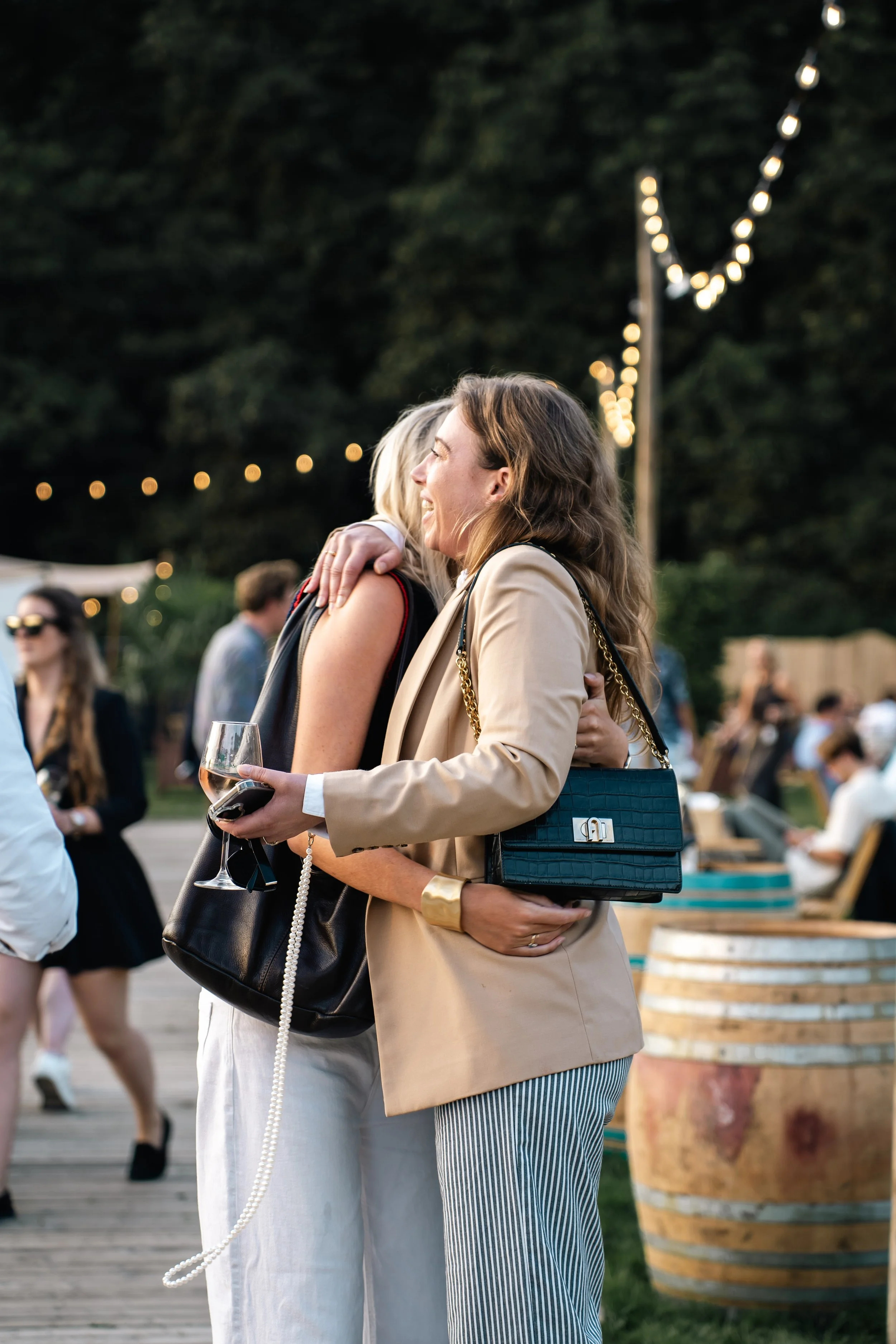 Twee vrouwen die elkaar omarmen en lachen op een buitenfeest, met achtergrond van mensen, boomverlichting en wijn.