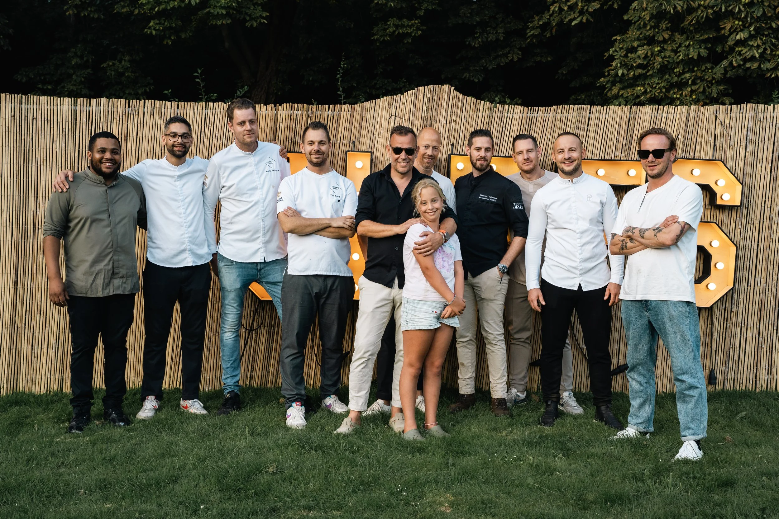 Groep mensen poseert voor een houten omheining, met een versiering van grote letters met lampjes op de achtergrond.