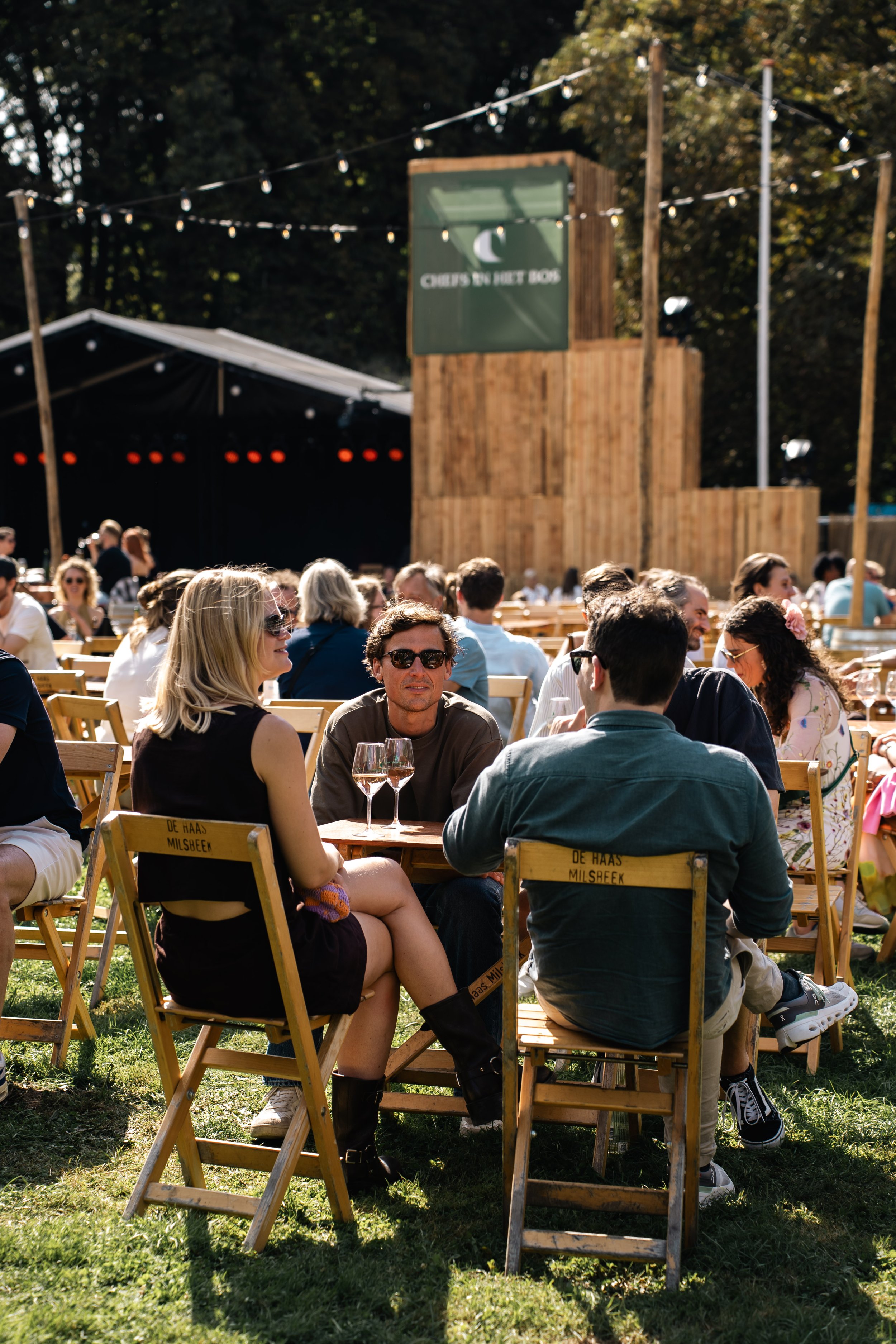Mensen zitten aan houten picknicktafels op een buitenfestival, met een podium, verlichting en bos op de achtergrond.