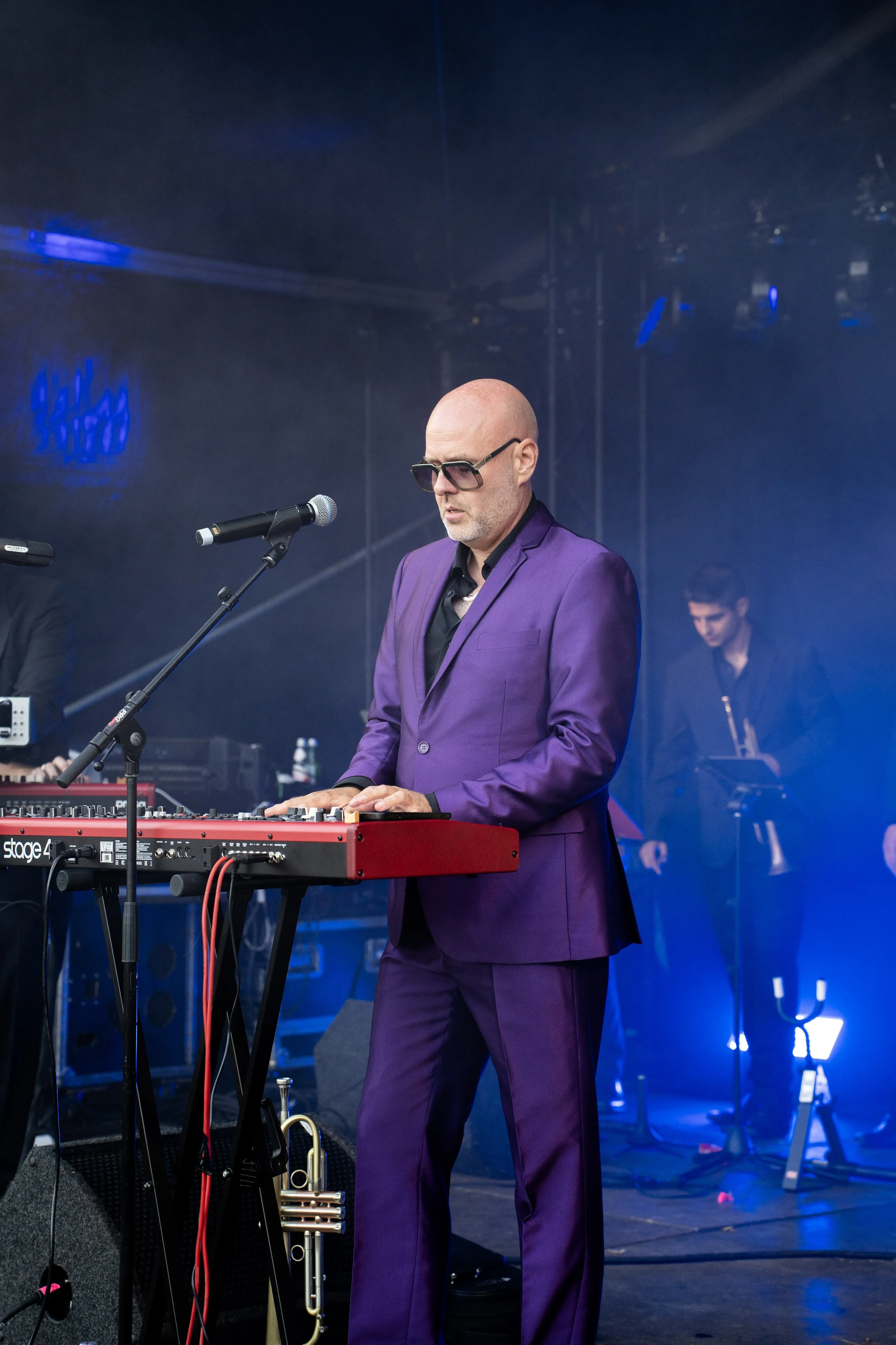 Man in een paarse pak en donkere zonnebril speelt keyboard op een podium, met een microfoon voor hem en muziekinstrumenten op de achtergrond, op een podium met blauwe verlichting.