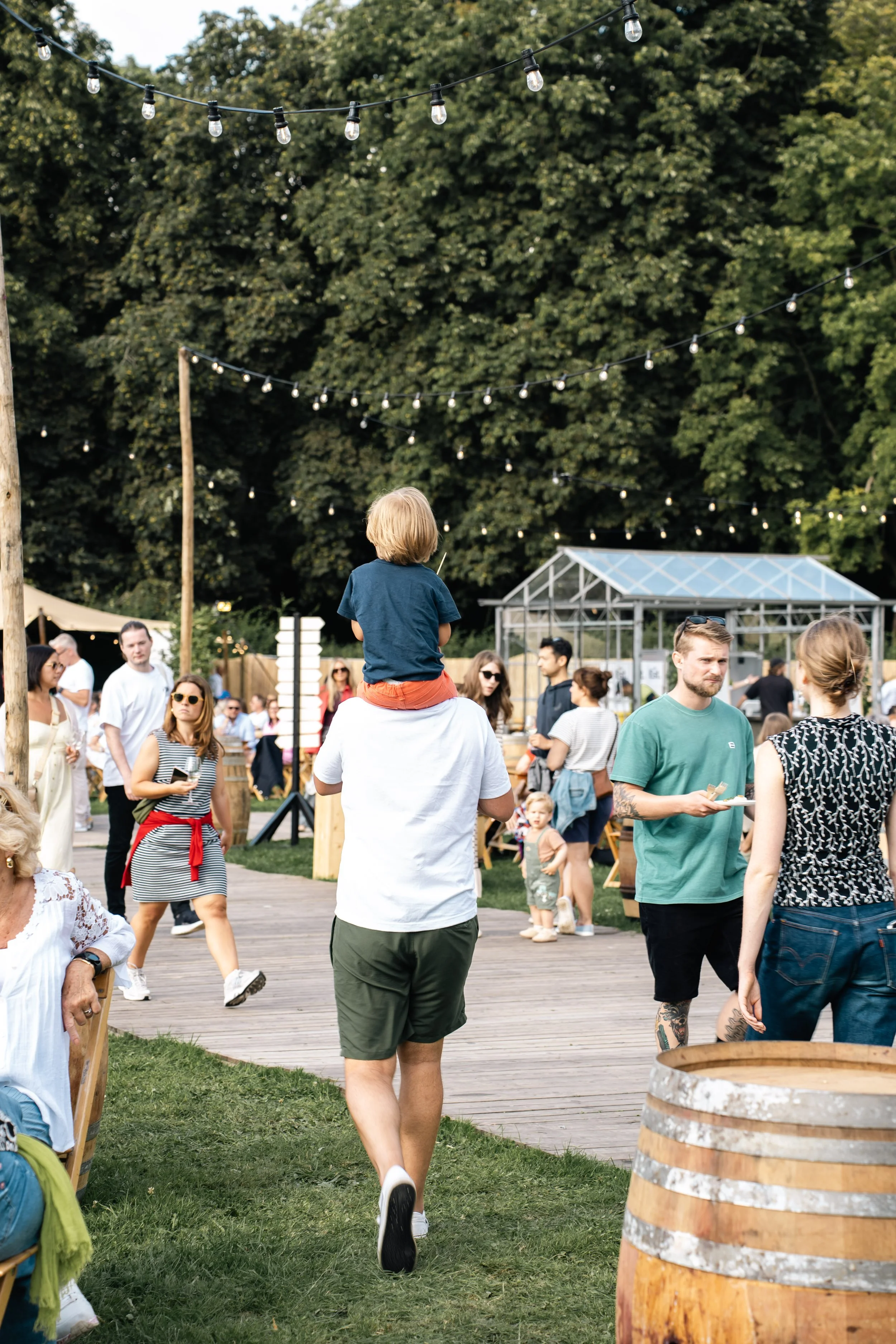 Mensen op een buitenevenement met bomen en lichtslingers, een kind op de schouders van een man