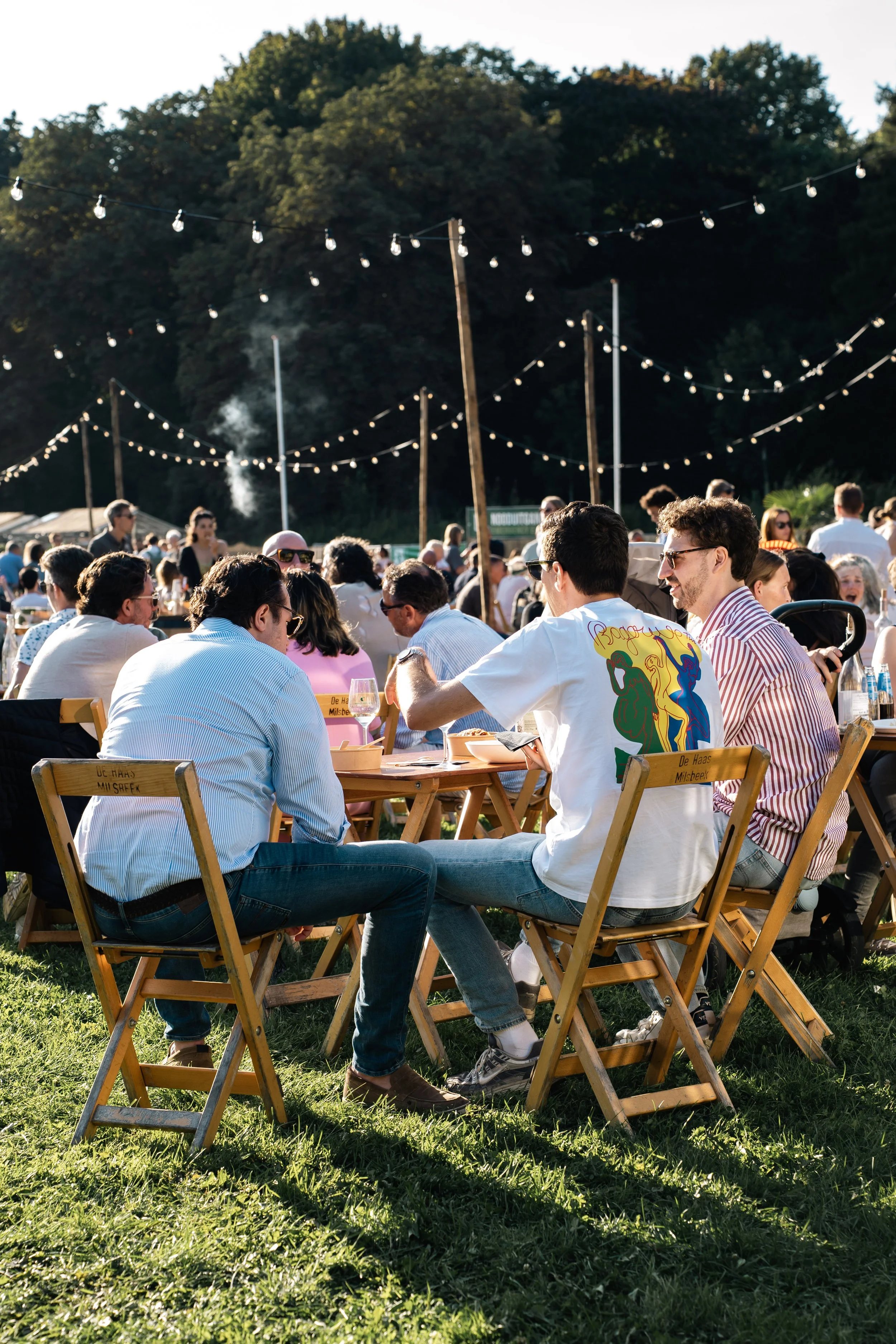 Mensen zitten en praten aan tafels op een openlucht festival of evenement, met stringlichten boven hen en een bosachtige achtergrond.