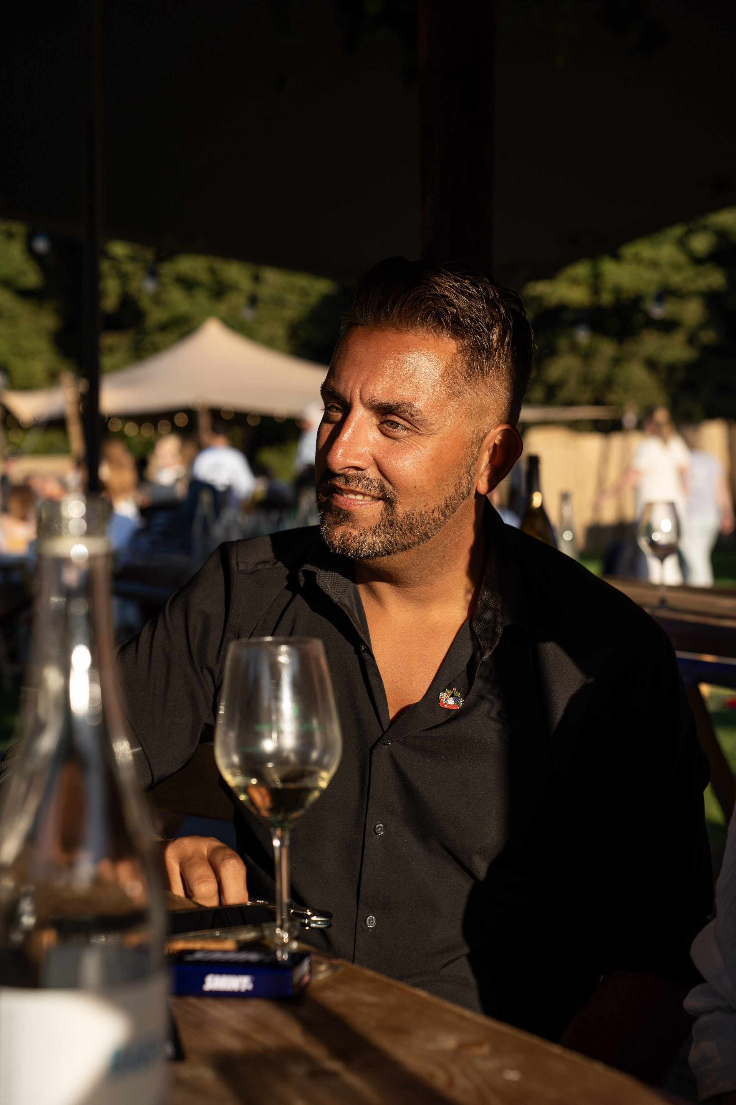 Man met korte donker haar en een baard glimlacht, zittend aan een tafel in een buitenrestaurant met witte tenten en groene bomen op de achtergrond, in de avond met warm zonlicht.
