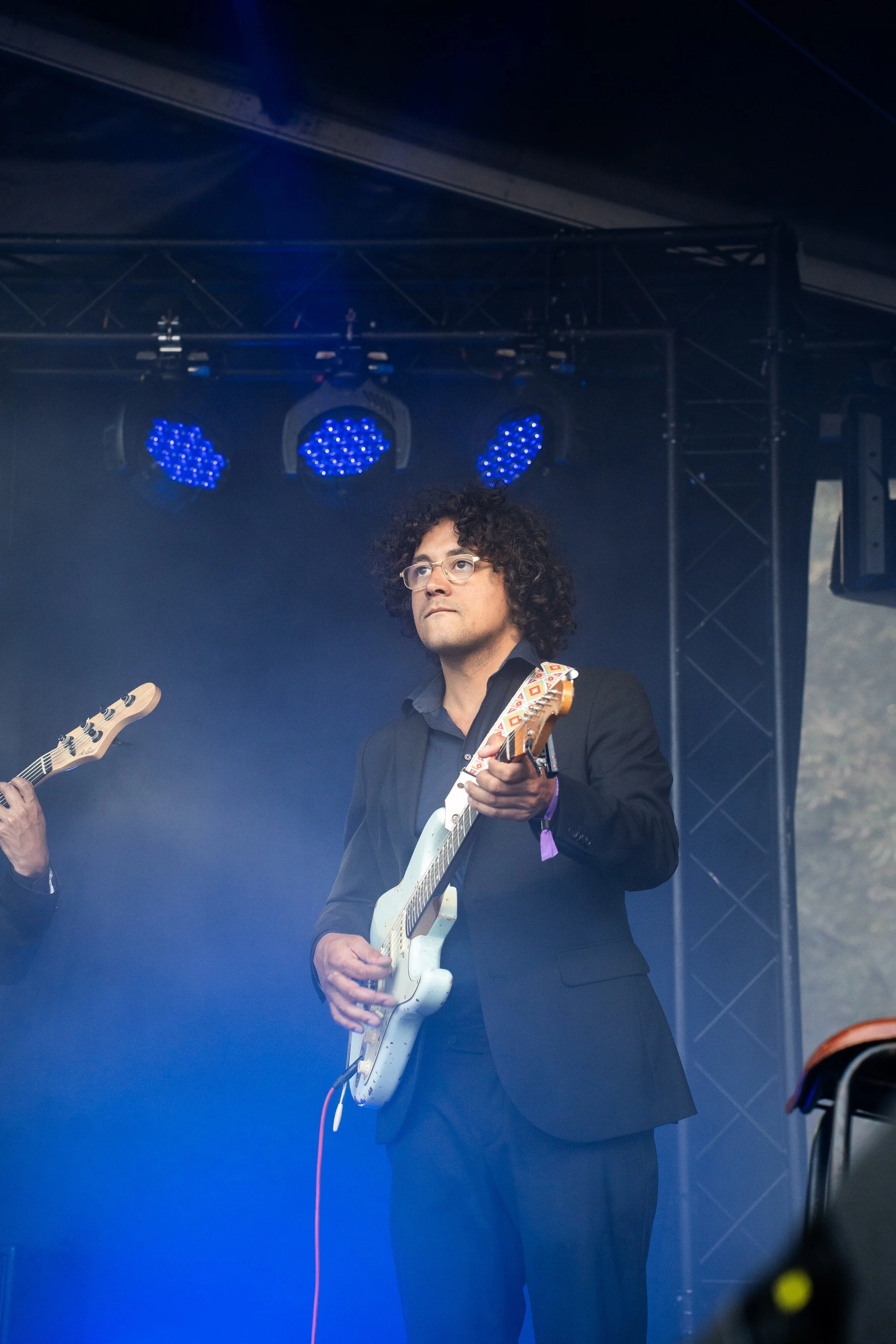 Man met bril en zwart pak speelt elektrische gitaar op een podium met blauwe en paarse verlichting.