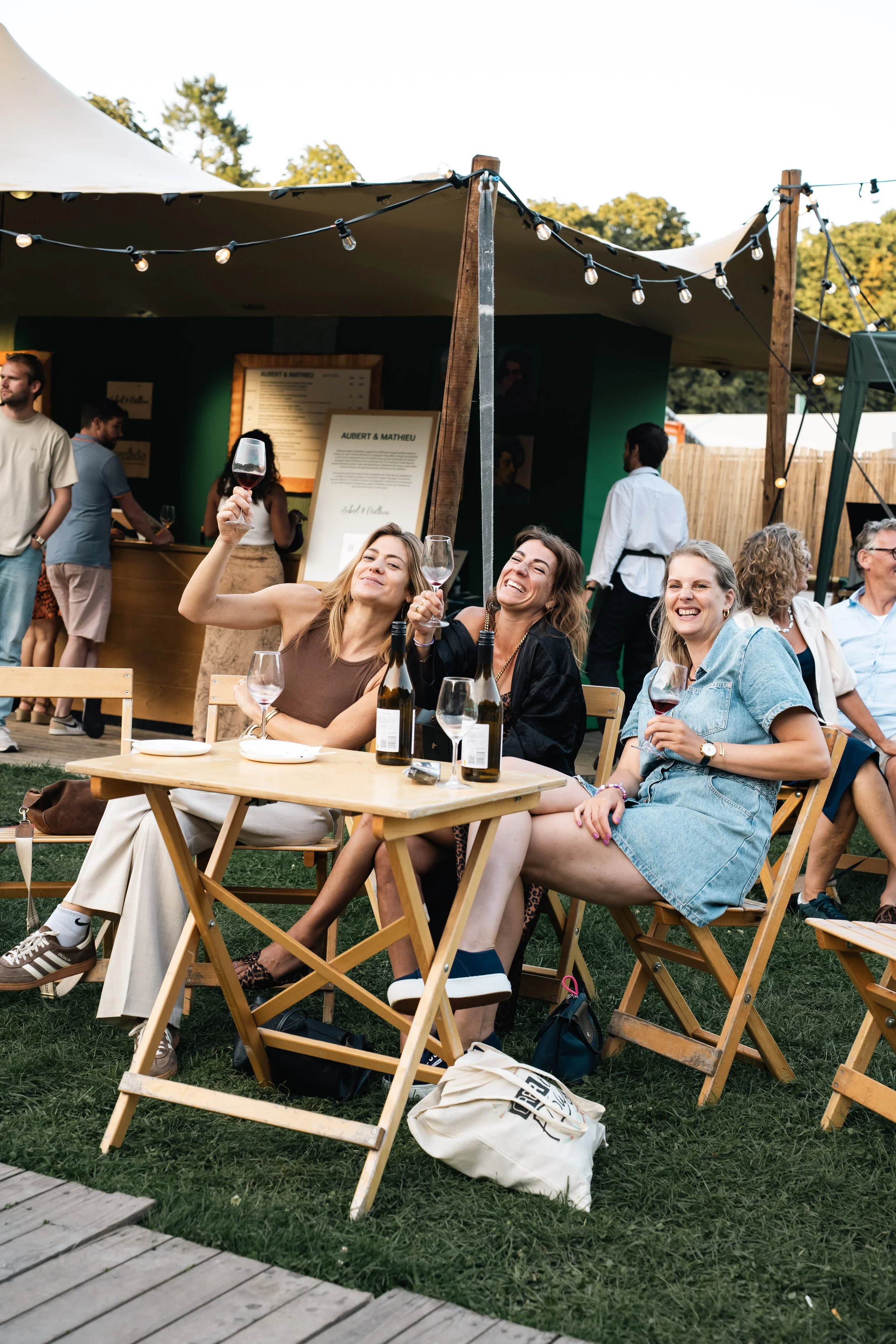 Groepen mensen genieten van een buitenevenement onder een tent met feestverlichting. Drie vrouwen zitten aan een houten tafel met wijn, lachend en proostend. Achter hen staan mensen in de rij bij een bar, terwijl anderen in gesprek zijn of op een ter