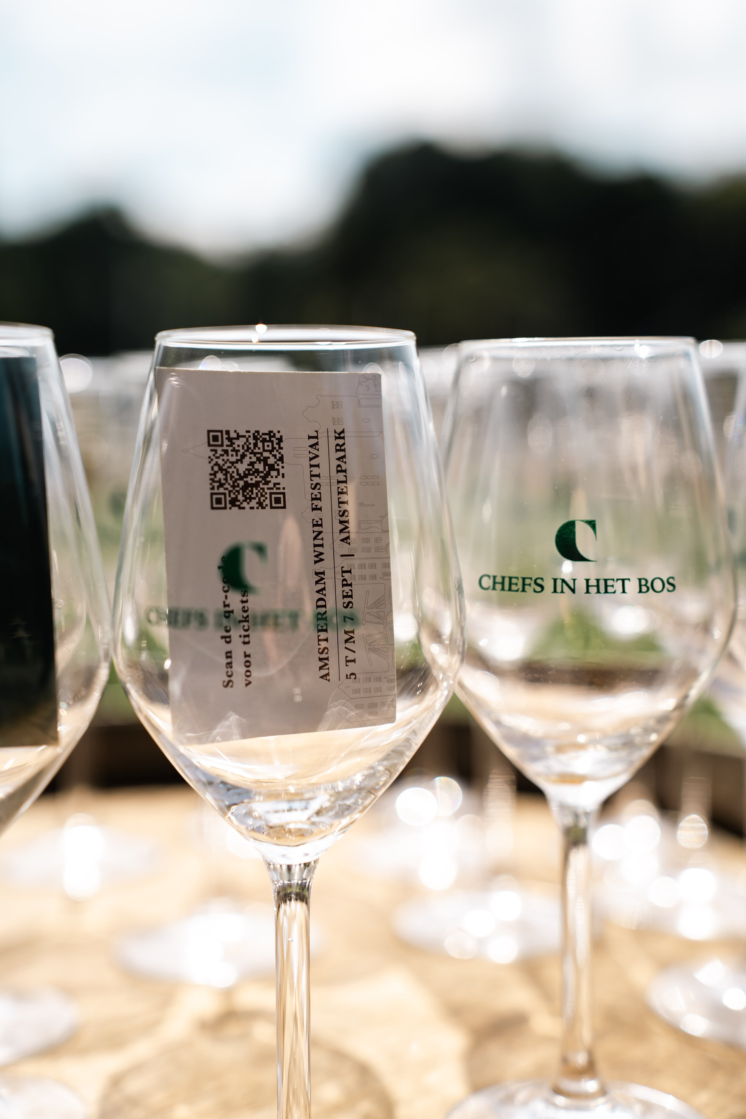 Verschillende lege wijn- of champagneglazen op een tafel met een ticket of programmalabel in één glas, achtergrond met natuurlijke buitenlucht.