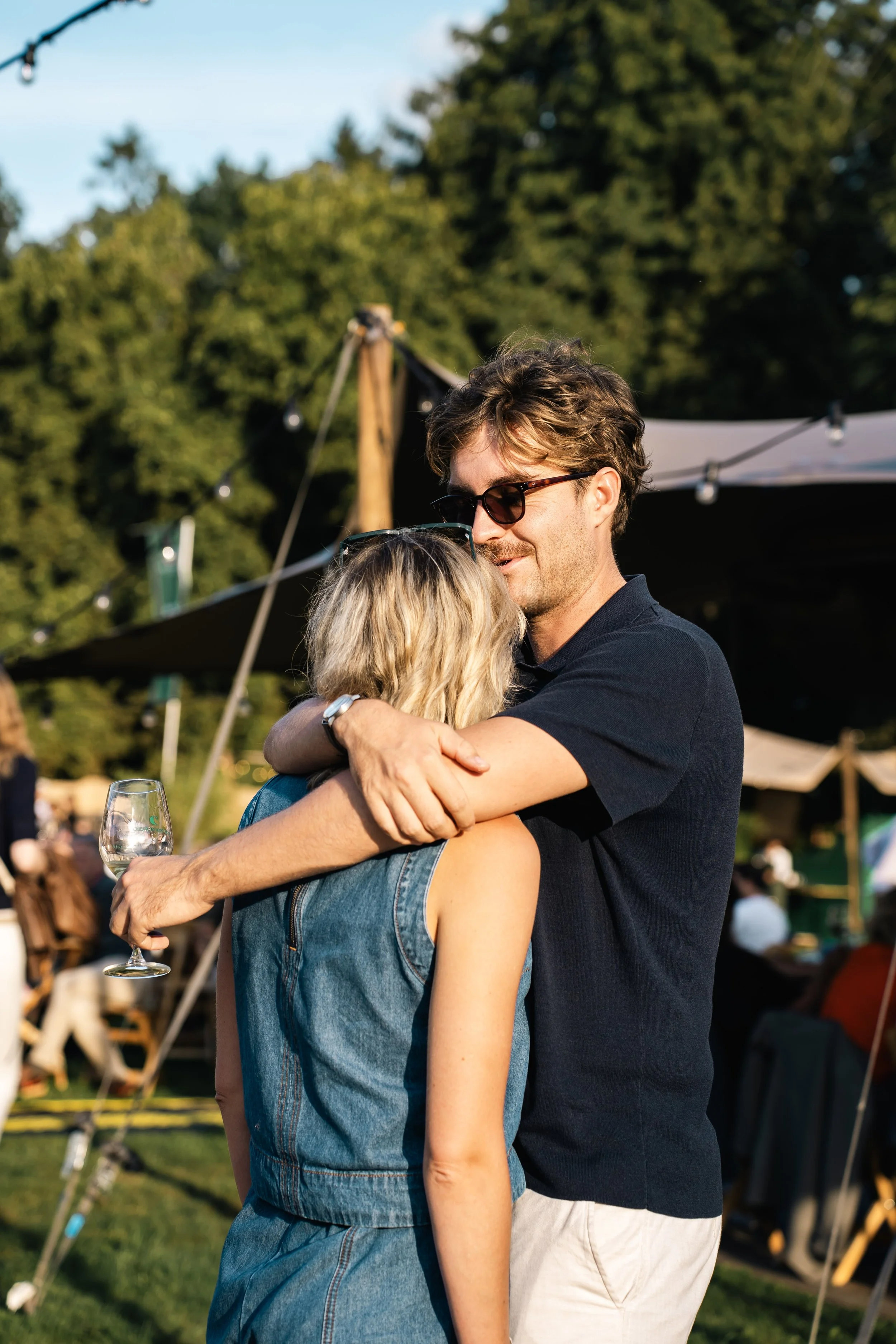 Man en vrouw omhelzen elkaar op een feestje in de buitenlucht, man draagt een zwarte polo en witte pantalon, vrouw draagt een denim jas, man draagt een zonnebril.