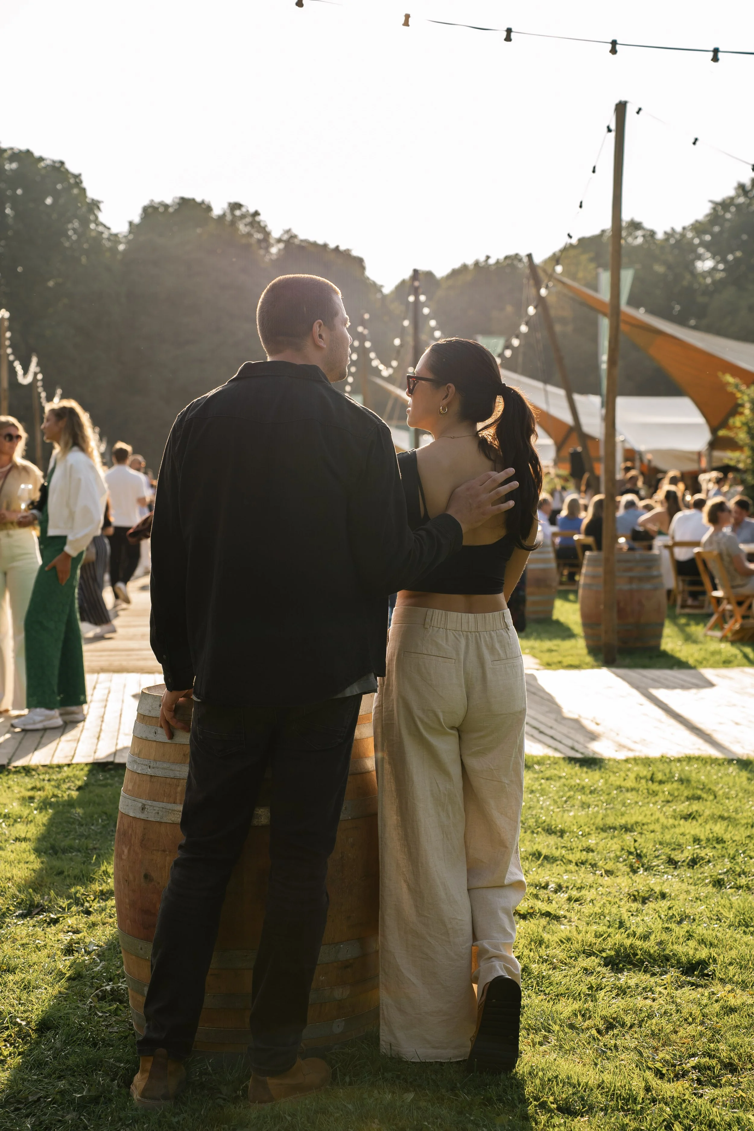 Twee mensen staan dicht bij elkaar in een openluchtfeest, terwijl de zon ondergaat. Mensen dansen en sociaal samen zijn op de achtergrond, met tenten en lichtslingers.