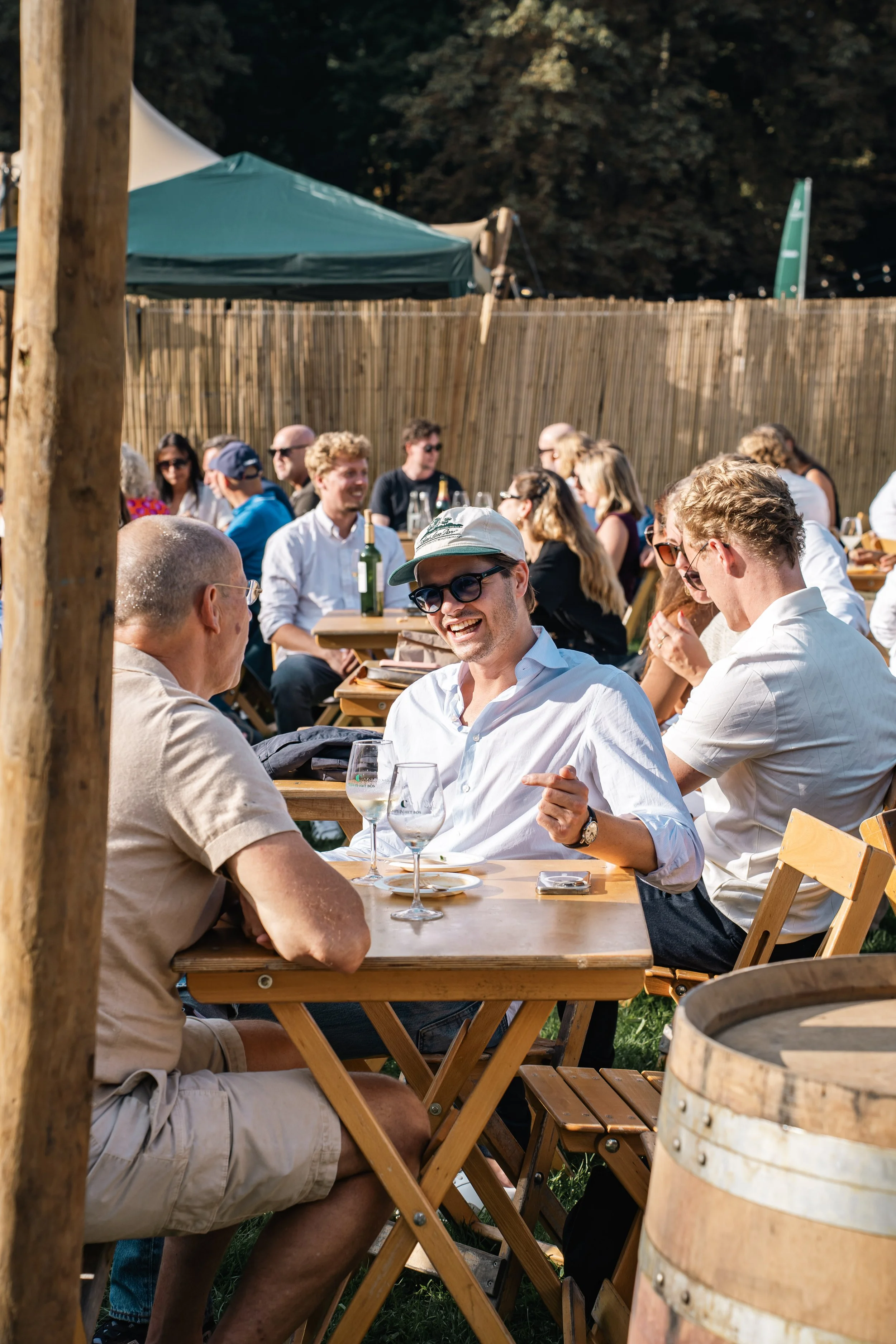 Mensen genieten samen in een tuin of terras, buiten onder een blauwe en groene tent, met houten en wijnfles op tafel, omgeven door een houten en bamboe hek.