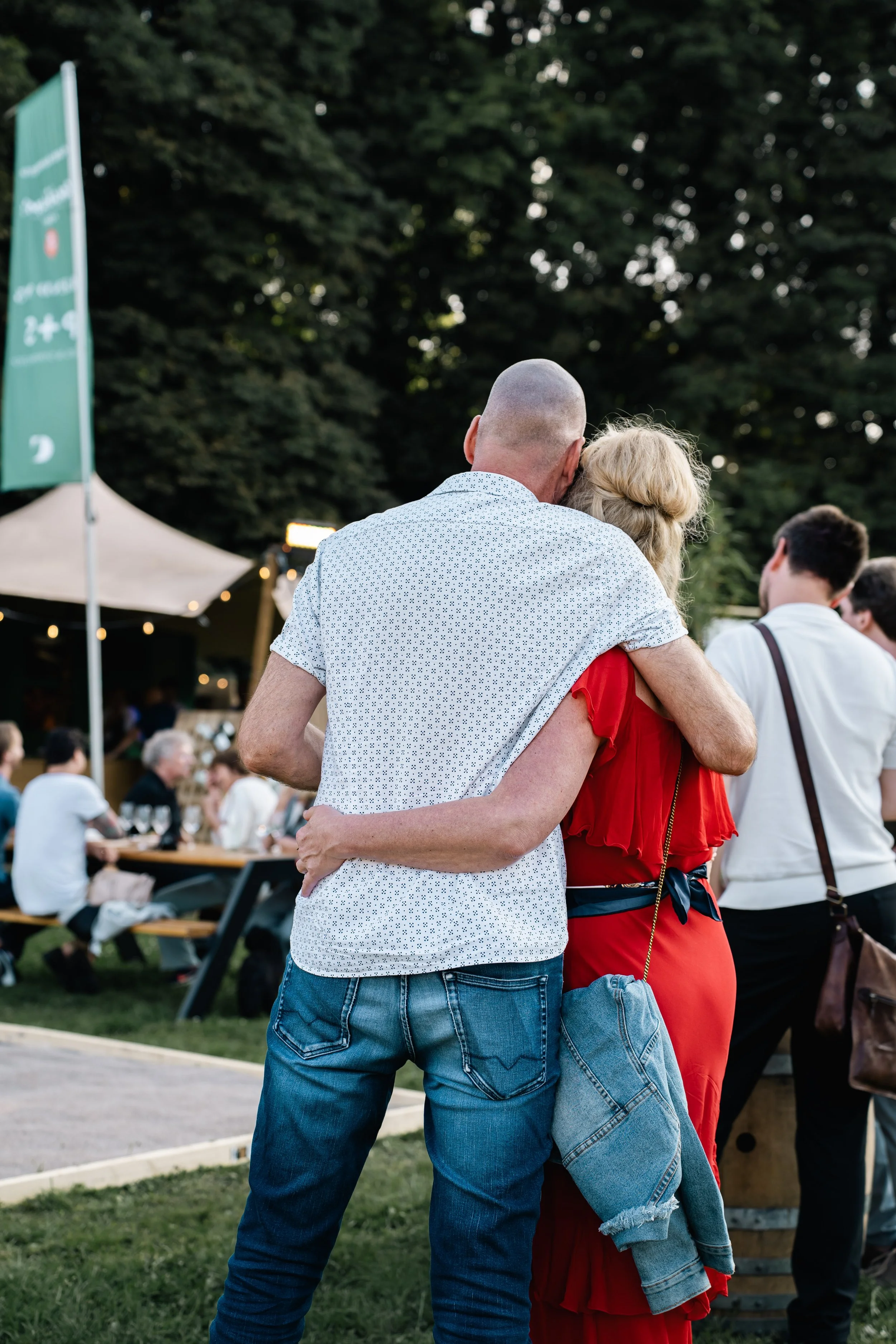 Een paar omhelst elkaar op een buitenfestival. Ze staan met de rug naar de camera, omringd door andere mensen en een foodtruck op de achtergrond.