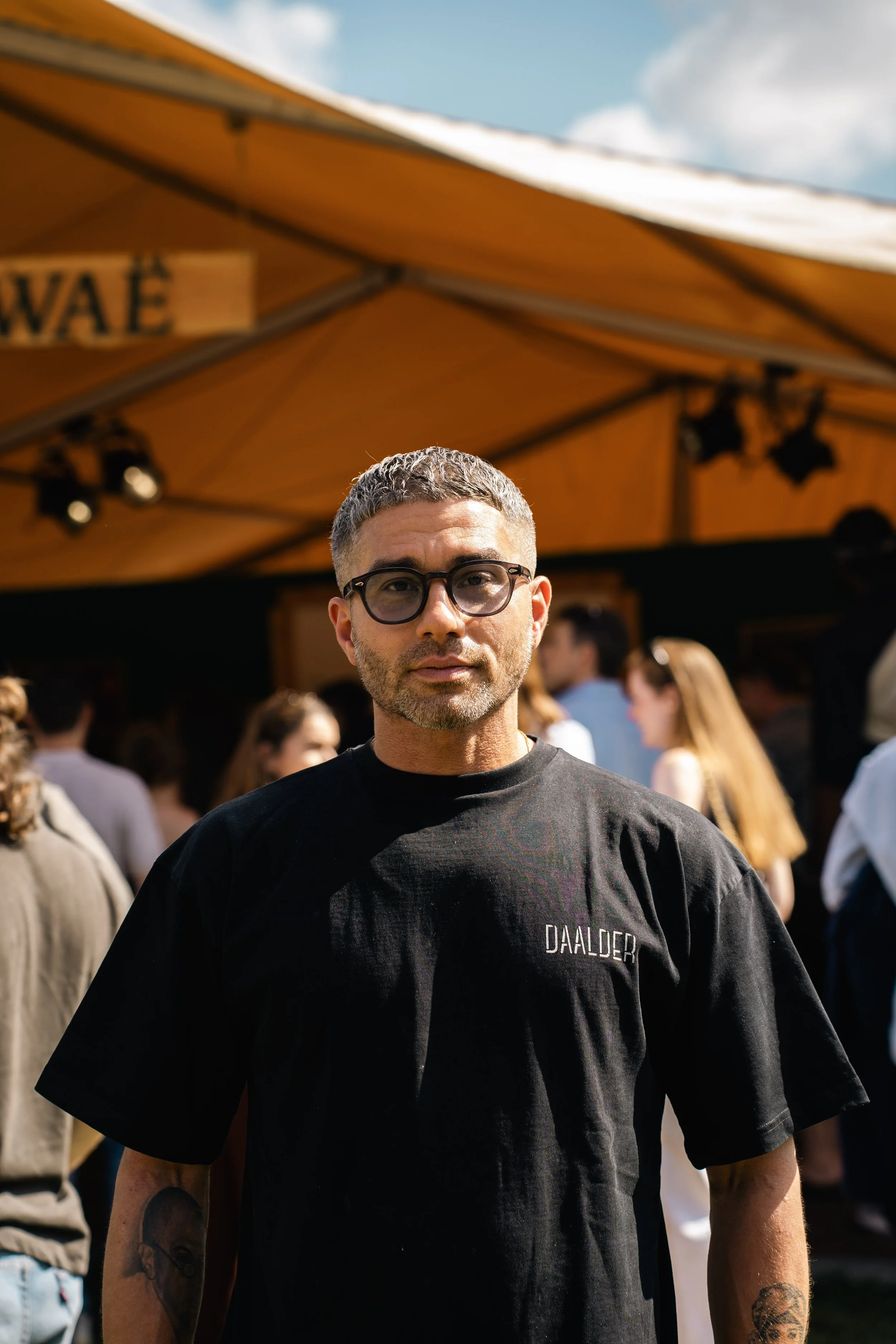 een man met korte grijze haar en brillen draagt een zwart T-shirt met de tekst 'DALDER' erop en staat in een drukke buitenomgeving met mensen en een oranje tent op de achtergrond.