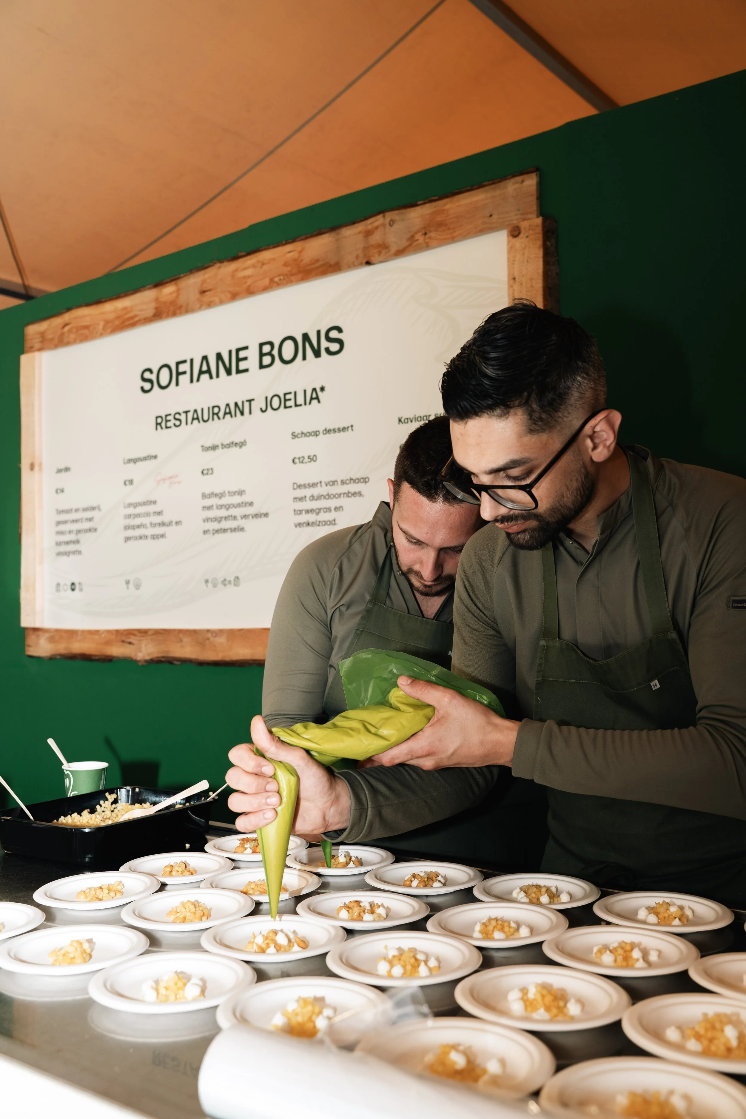 Twee koks in uniform bereiden en garneren kleine gerechten in een professionele keuken. Ze plaatsen de gerechten op een tafel met witte doeken. Op de achtergrond een bord met de naam Sofiane Bons en een menu.