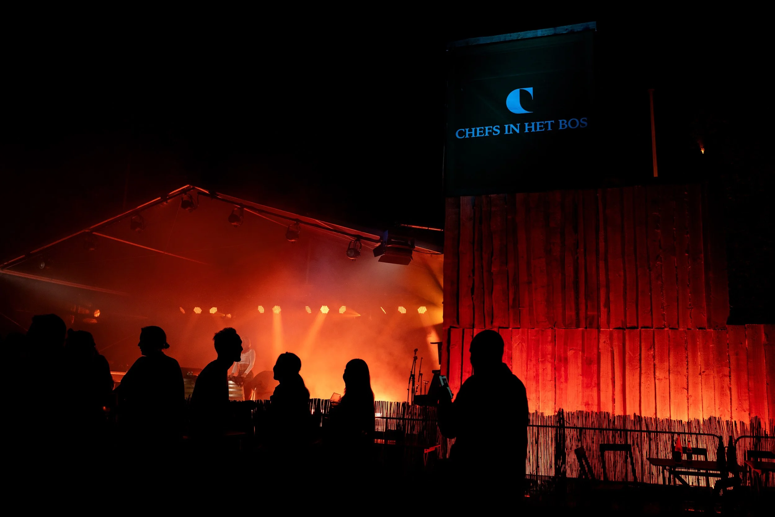 Een donkeravondbeeld met silhouetten van mensen die naar een podium kijken, verlicht door rode en oranje lichten.
