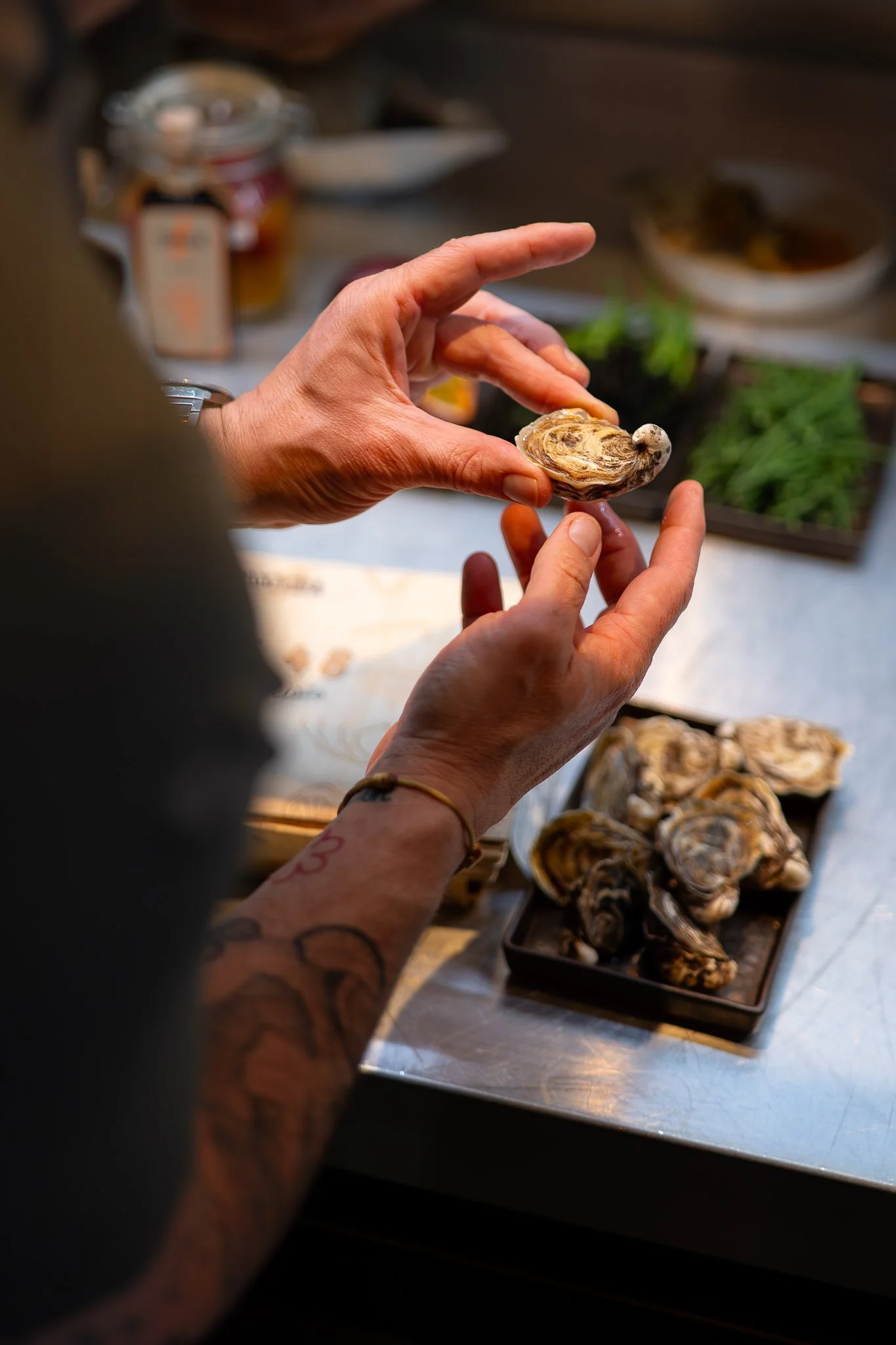 Persoon die een oester vasthoudt in een keuken, met schaaltjes met oesters op een tafel op de achtergrond.