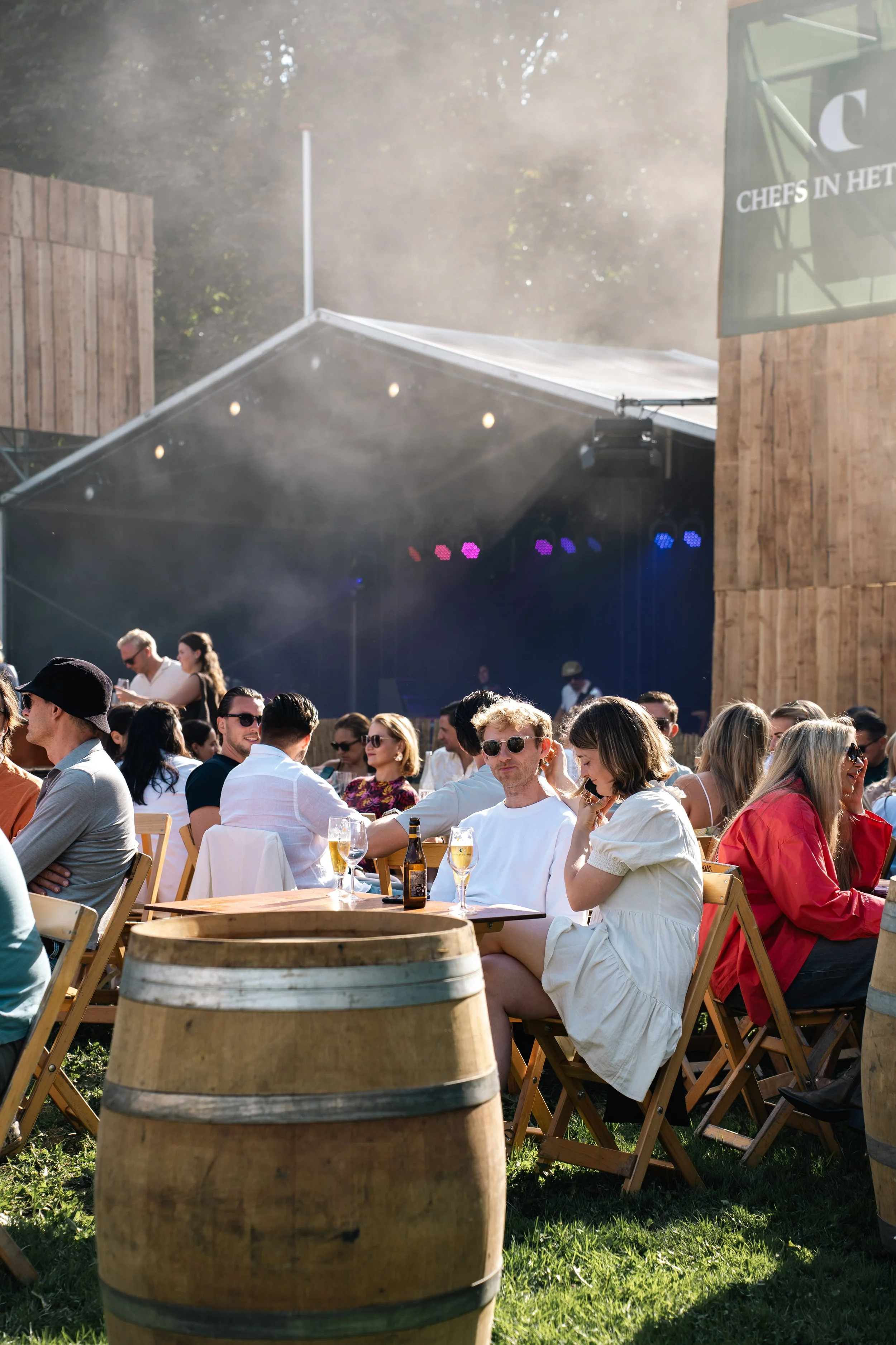Een groep mensen die buiten aan een feestje of evenement deelnemen, zittend aan houten tafels en stoelen onder een tent. In de achtergrond is een houten gebouw en een groot groene vlag zichtbaar, met mensen die genieten en drinken.