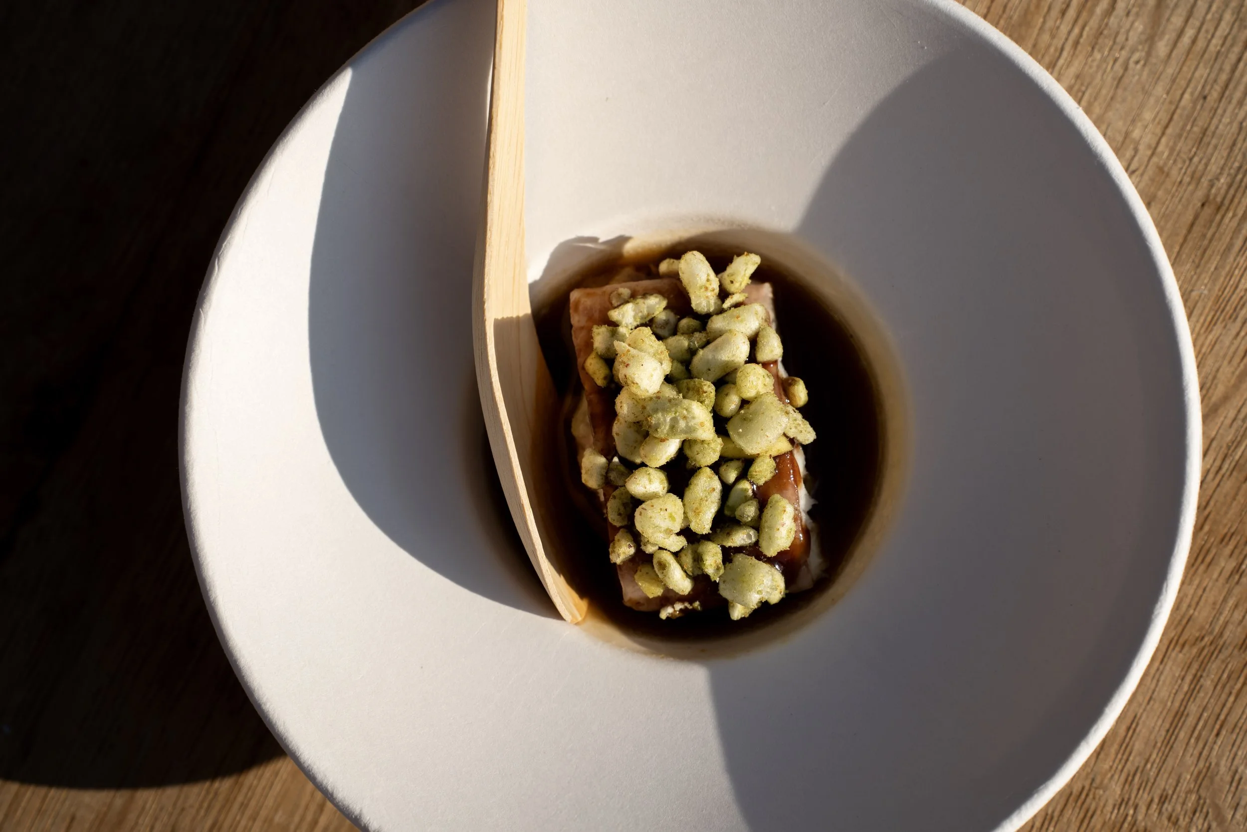 Een wit bord met daarop een houten lepeltje dat een stukje vlees met horensoep bedekt en bedekt is met knapperige pinda's of cashewnoten op een houten tafel.