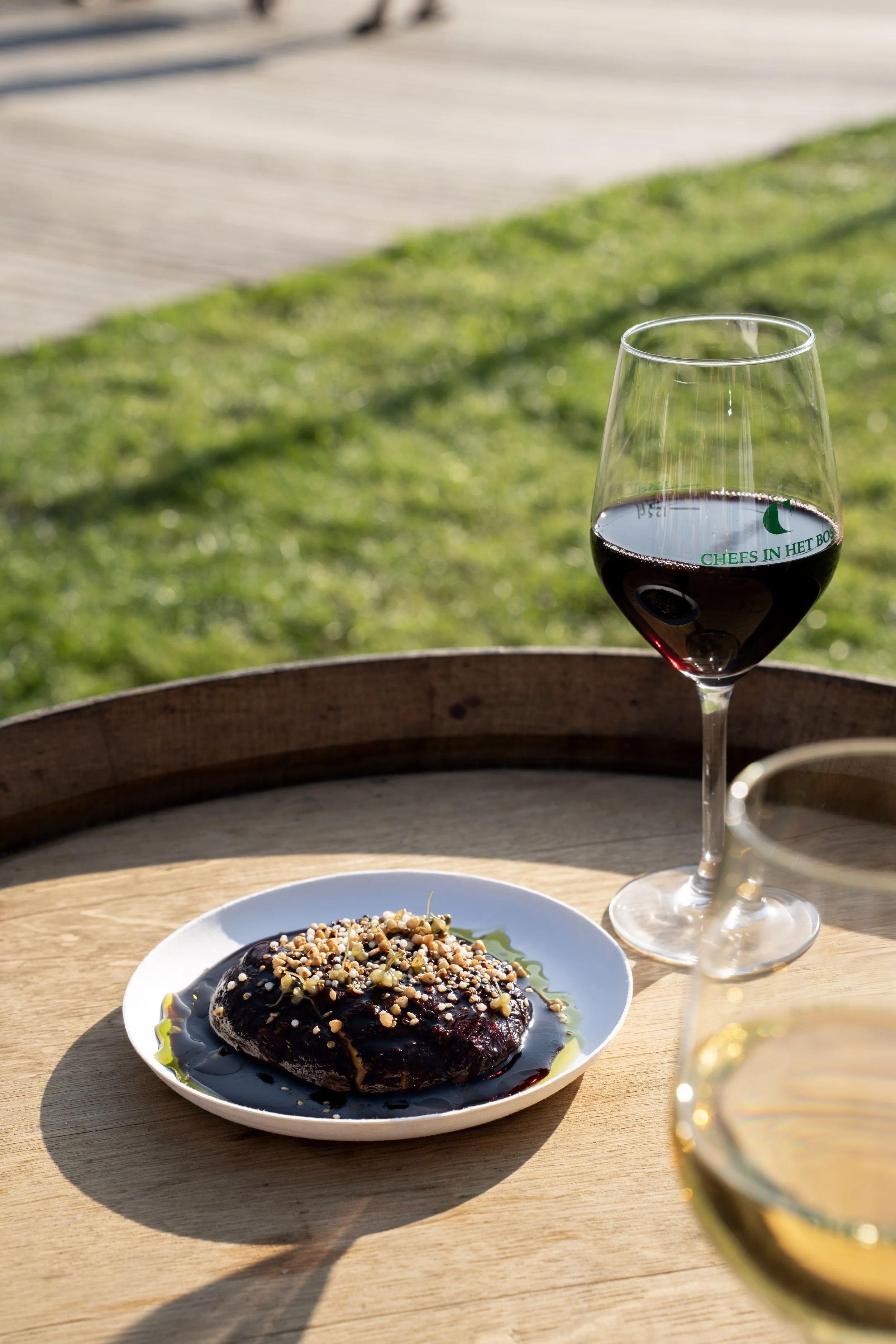 Een glas rode wijn op een houten tafel met daarop ook een bord met een dessert bedekt met chocolade en noten, en een tweede glas witte wijn op de achtergrond. Op de achtergrond is gras en een terras zichtbaar.