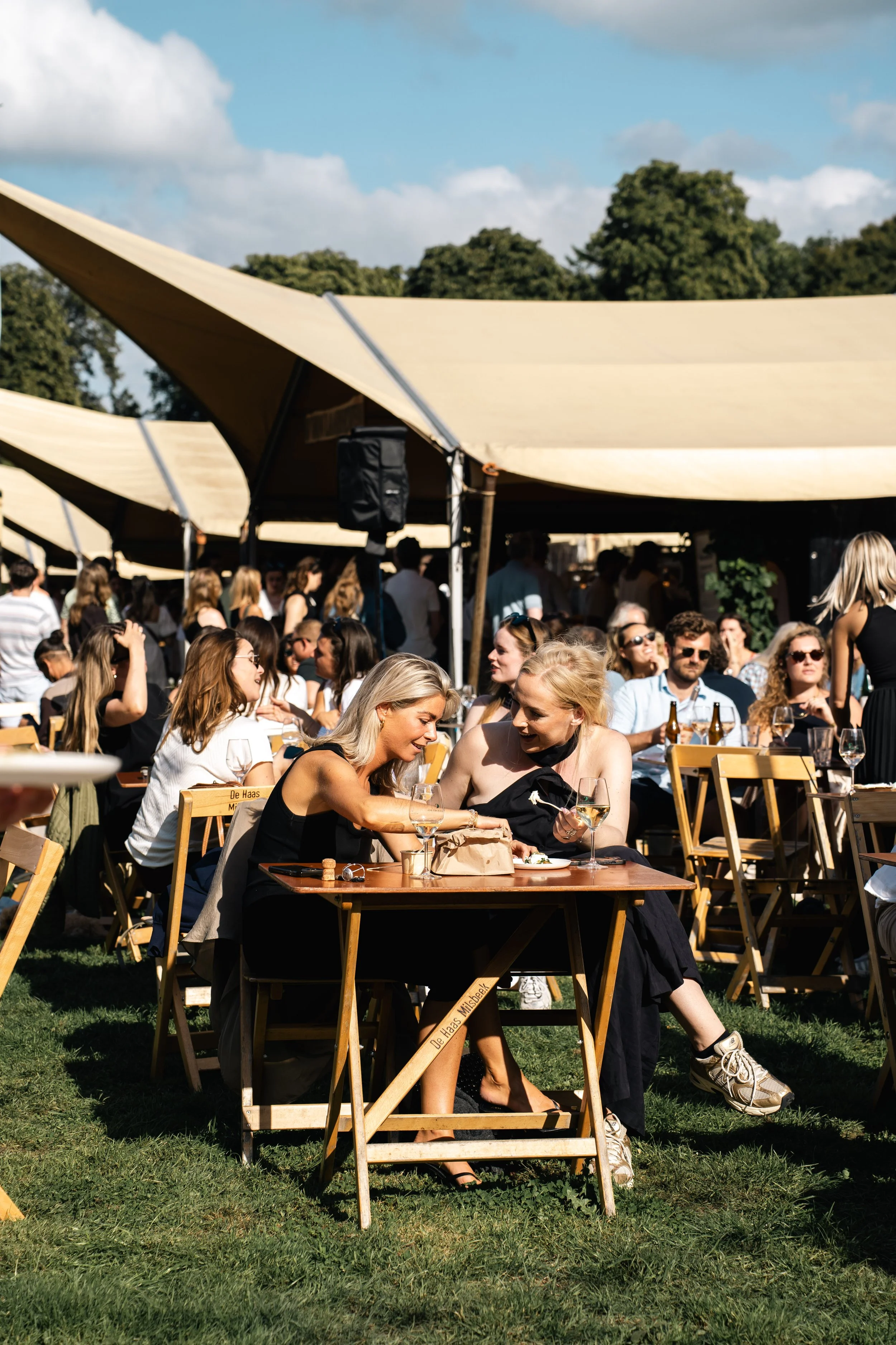 Een drukke buiten eetgelegenheid met mensen die genieten van eten en drinken onder grote beige parasols op een grasveld, met bomen en een blauwe lucht met wolken op de achtergrond.