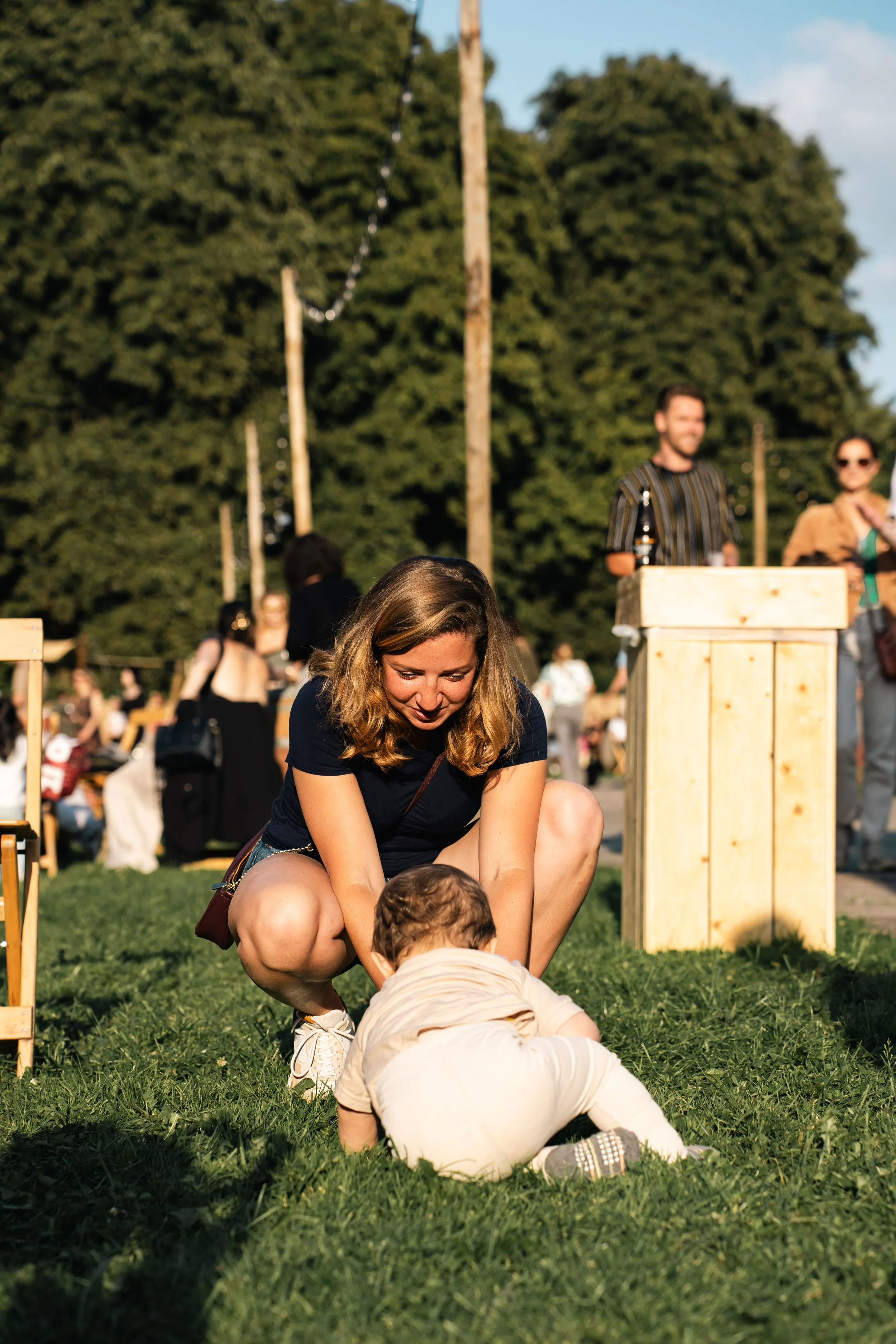Vrouw knielend op gras, spelend met baby in park tijdens feestje met andere mensen op achtergrond.