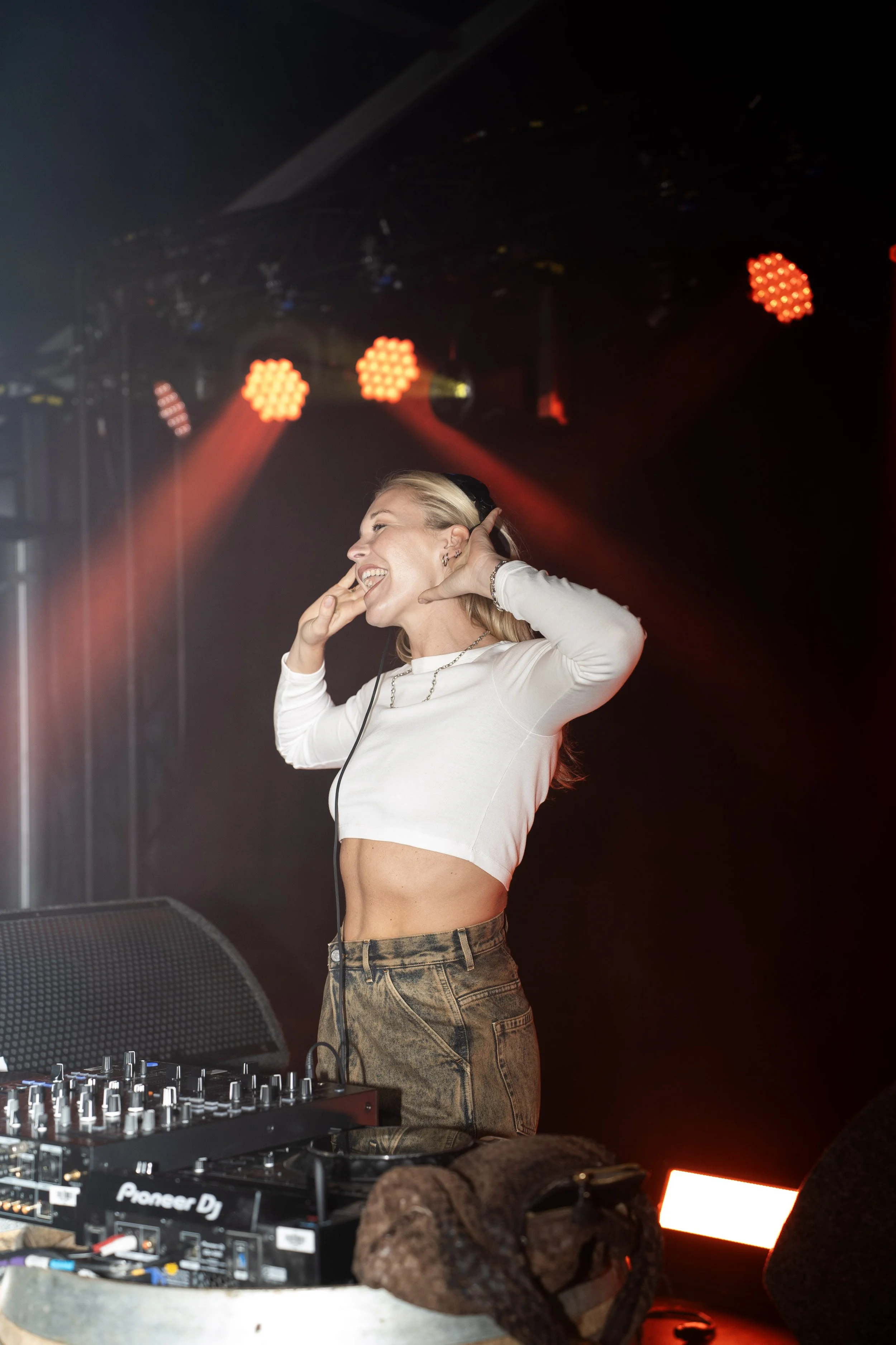 Vrouw dj met lange blonde haar in een witte cropped top en camouflage broek draait aan haar hoofd, achter haar draait rode en gele licht bij een dj-set.