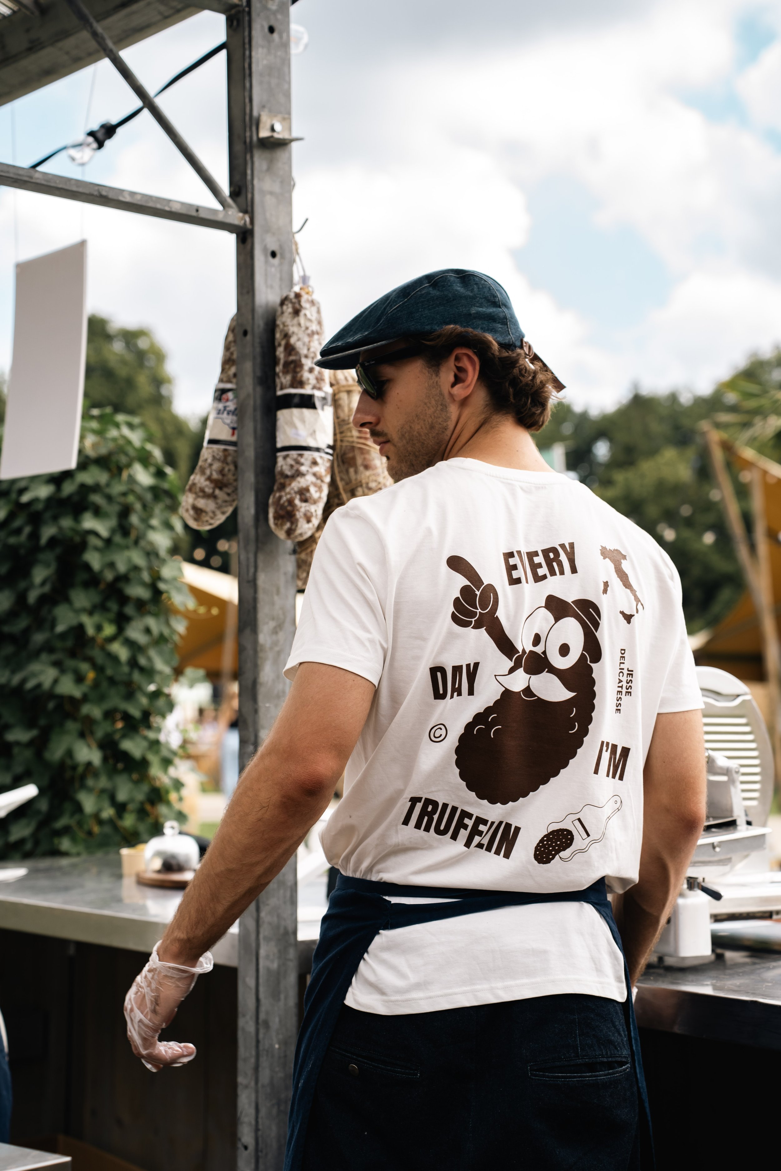 Man draagt een zwart schort, T-shirt met een grappige illustratie en tekst, vertelt of verkoopt worst of vleesproducten op een markt, naast een ijzeren frame met vleeswaren.