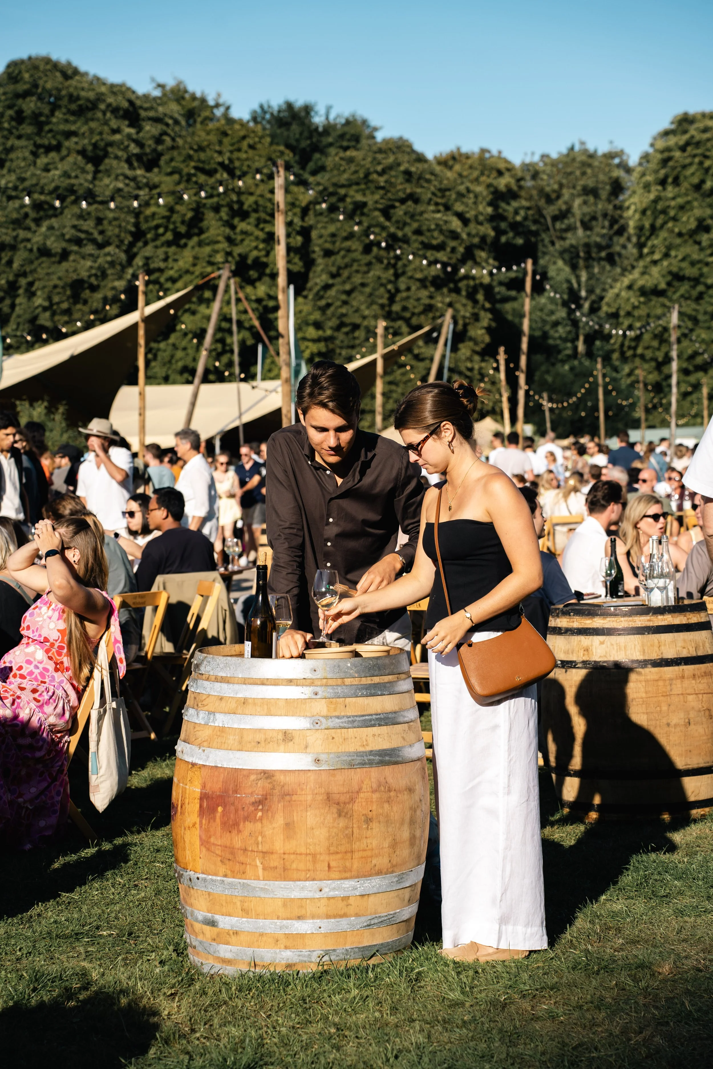 Een grote groep mensen bij een feest of festival op een grasveld, met tenten en lichtslingers, mensen zitten en staan in het zonlicht, twee mensen staan naast een wijnfles op een houten vat, de man serveert een glas wijn aan de vrouw.