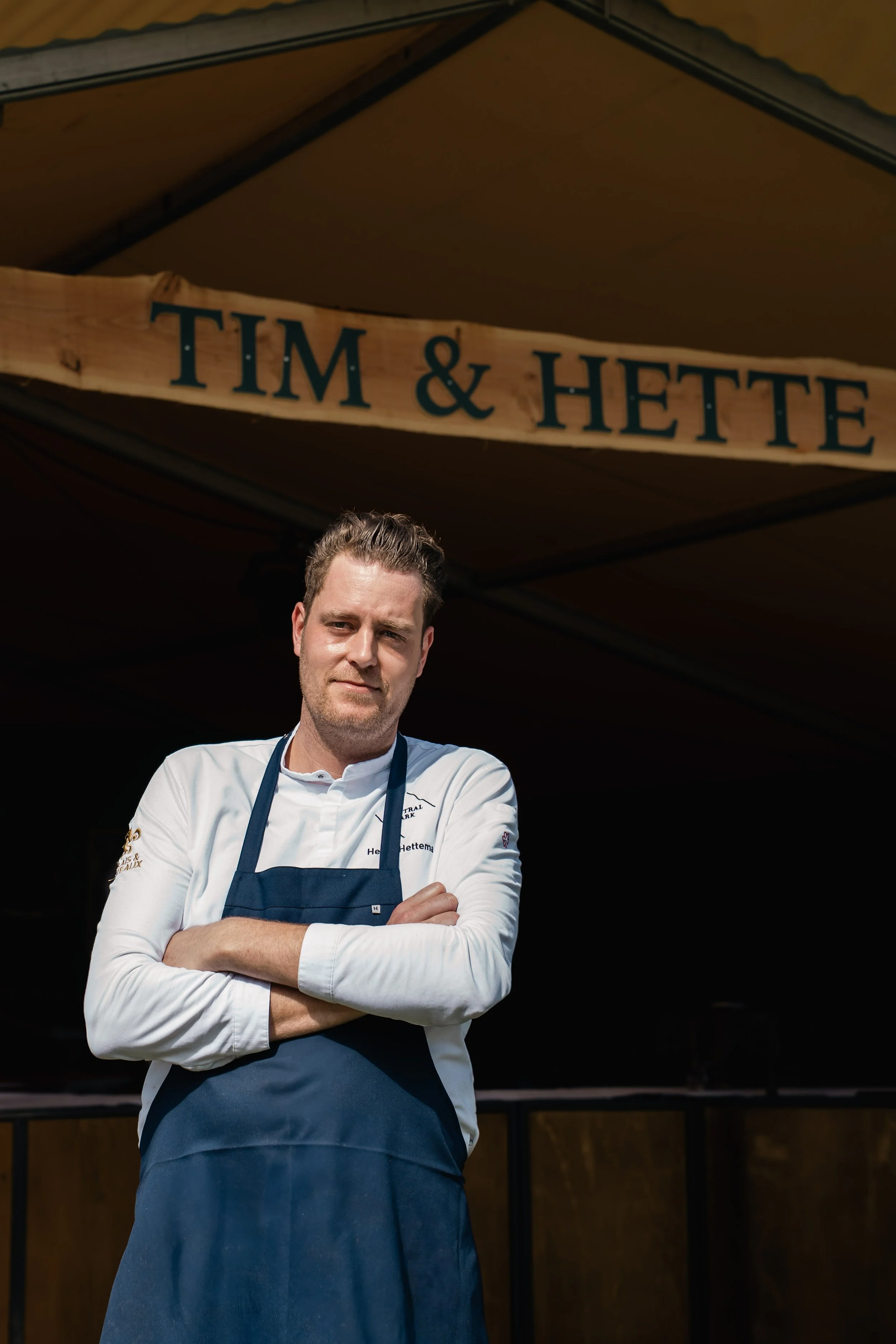 Een man in een witte chef-koksjas en een donkerblauwe schort staat met gekruiste armen onder een houten uithangbord met de tekst 'TIM & HETTE'.